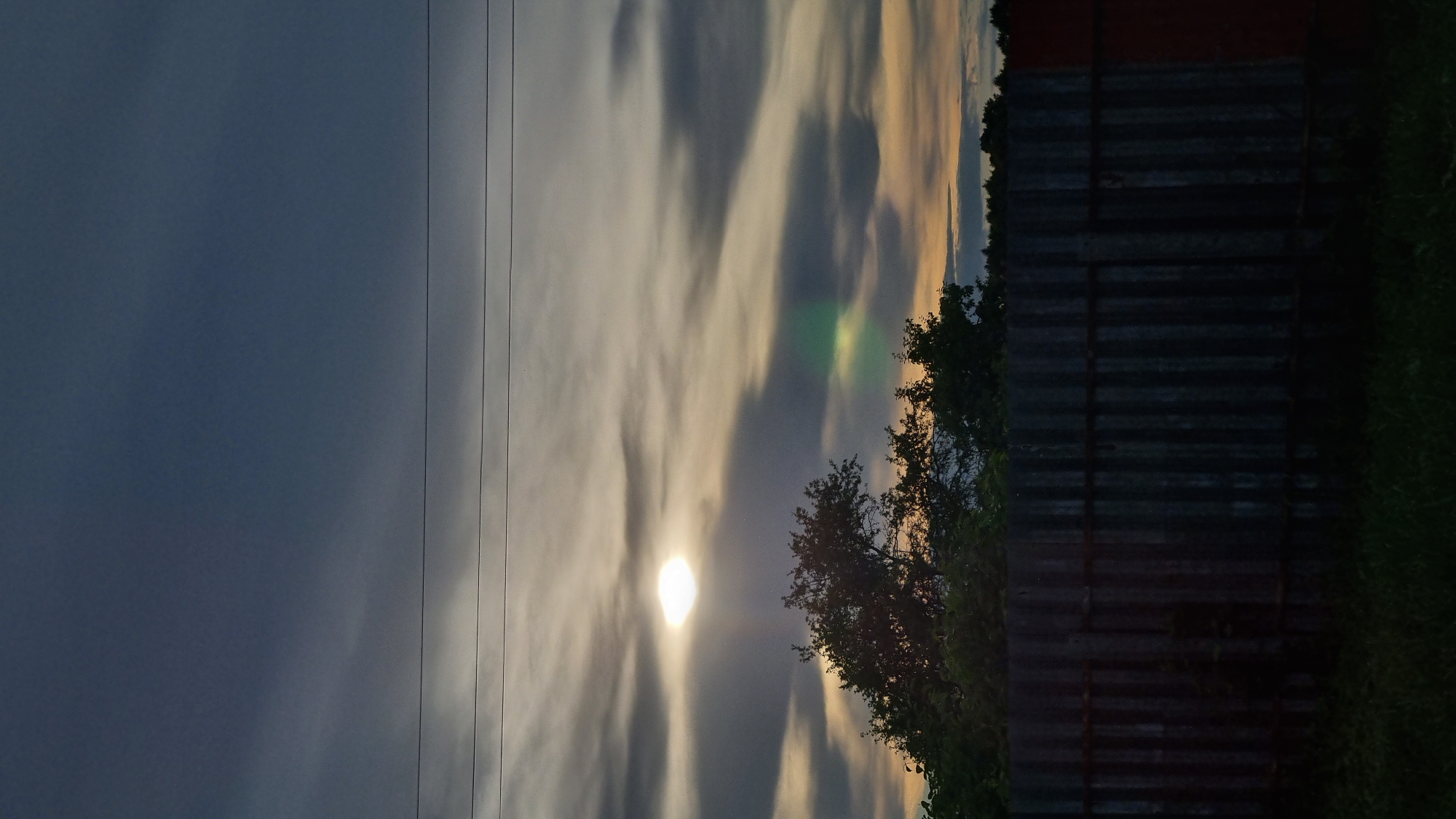 A vertical, medium shot captures a twilight sky above a corrugated metal fence in Vysokoe, Russia. The sky transitions from a deep blue at the top to lighter shades of gray and yellow towards the horizon, with wispy clouds creating streaks across the scene. A bright, circular object, likely the sun, is partially obscured by clouds in the upper left quadrant. A faint green lens flare is visible in the lower right quadrant of the sky. Silhouetted trees form a dark line against the brighter sky at the horizon, behind the fence. The fence itself is dark and textured, with horizontal supports visible. In the foreground, dark green grass is visible. Two parallel, thin, dark horizontal lines, possibly power lines, traverse the upper portion of the image. There are no people or animals visible in the frame.