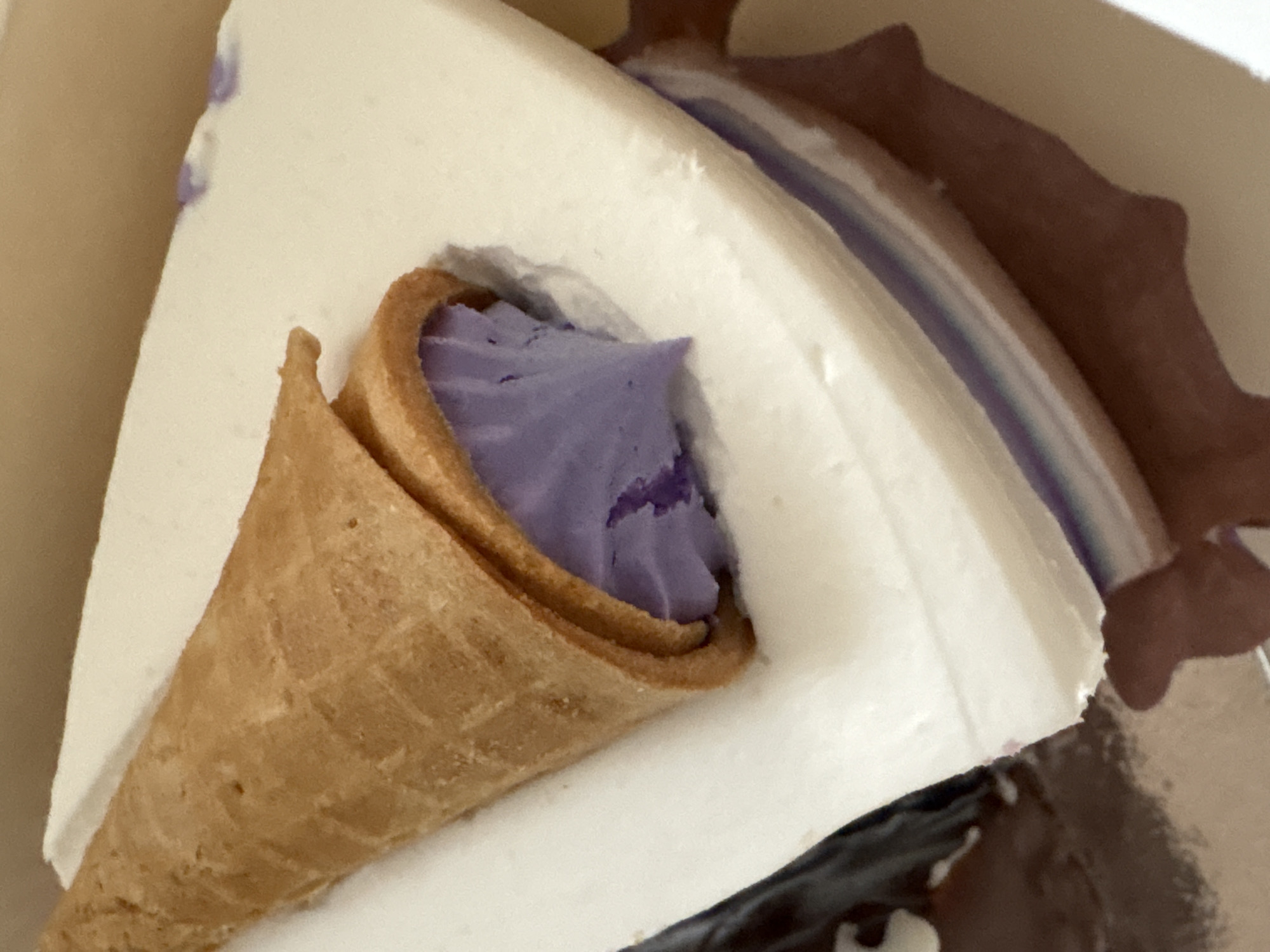 A close-up, high-angle shot captures a slice of dessert, likely a cake or ice cream cake, from above. The dessert features a thick, smooth white outer layer, resembling whipped cream, frosting, or soft-serve ice cream. Partially embedded into this white layer is a light brown, textured waffle cone. The cone is filled with a vibrant, swirled purple substance, possibly ube, taro, or a berry-flavored cream or ice cream.
A thin, horizontal stripe of the same purple color is visible within the main dessert slice, suggesting a layered construction. Dark brown elements, possibly chocolate cake layers or frosting, are visible in the background and at the bottom right of the frame, hinting at other components of the dessert. The overall composition suggests a rich and visually appealing treat. The lighting is direct, casting subtle shadows and highlighting the creamy texture of the white layer. There are no people or discernible text visible in the image.