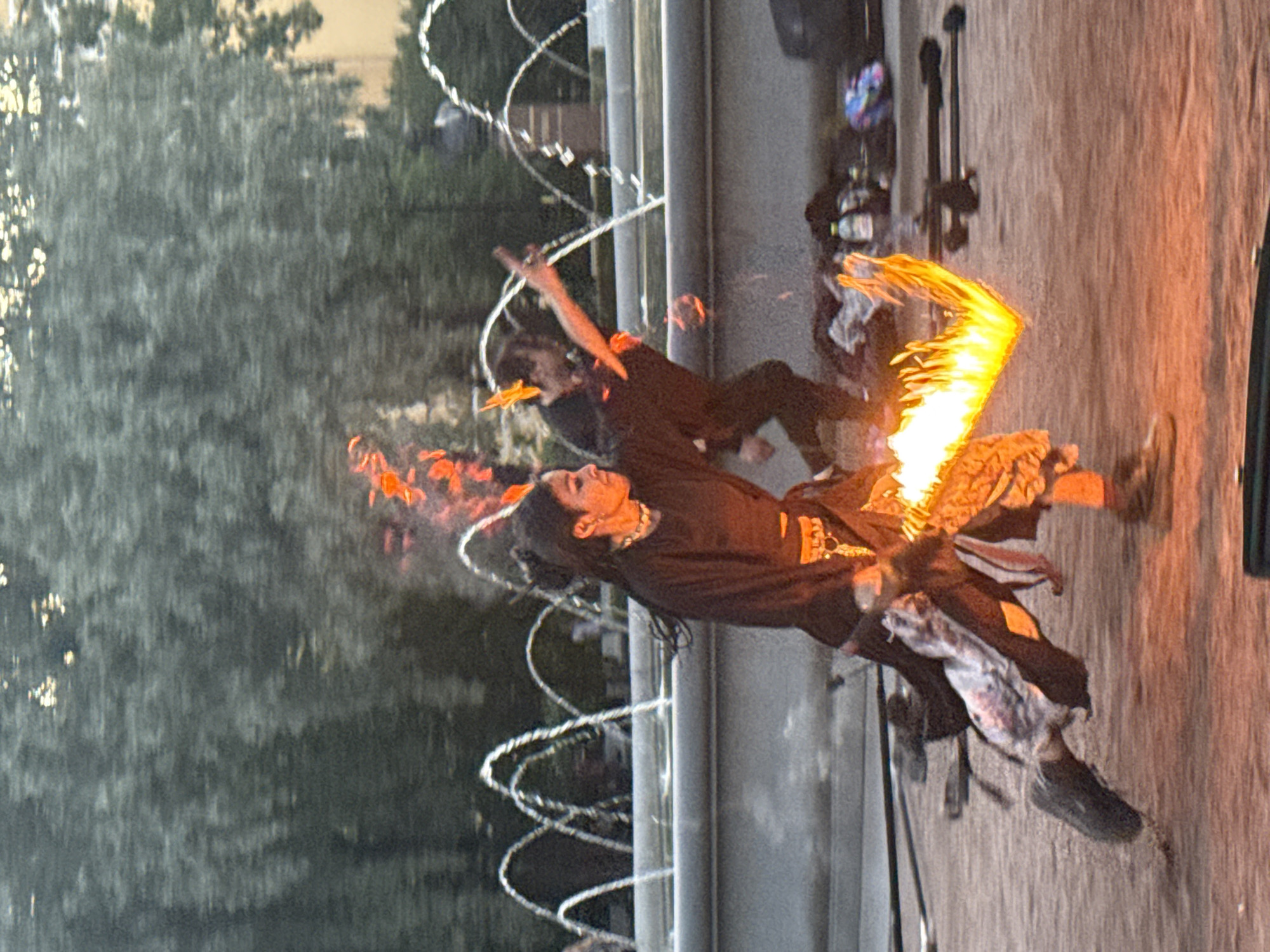 In Saint Petersburg, Russia, at dusk, two performers engage in a fire-dancing ritual near a tiered fountain. A woman, positioned in the foreground and to the left, executes a spinning motion with a flaming prop held in her right hand. She wears a black tunic and patterned trousers, with her hair pulled back in a bun. To her right, another performer, also in dark attire, appears to be in mid-action, possibly throwing or manipulating a fiery element. The background features the illuminated arcs of a fountain and the silhouettes of trees. The ground is a sandy or gravelly surface, with various objects, including bags and bottles, scattered around the performance area. The scene is characterized by the vibrant glow of fire against the dimming light.