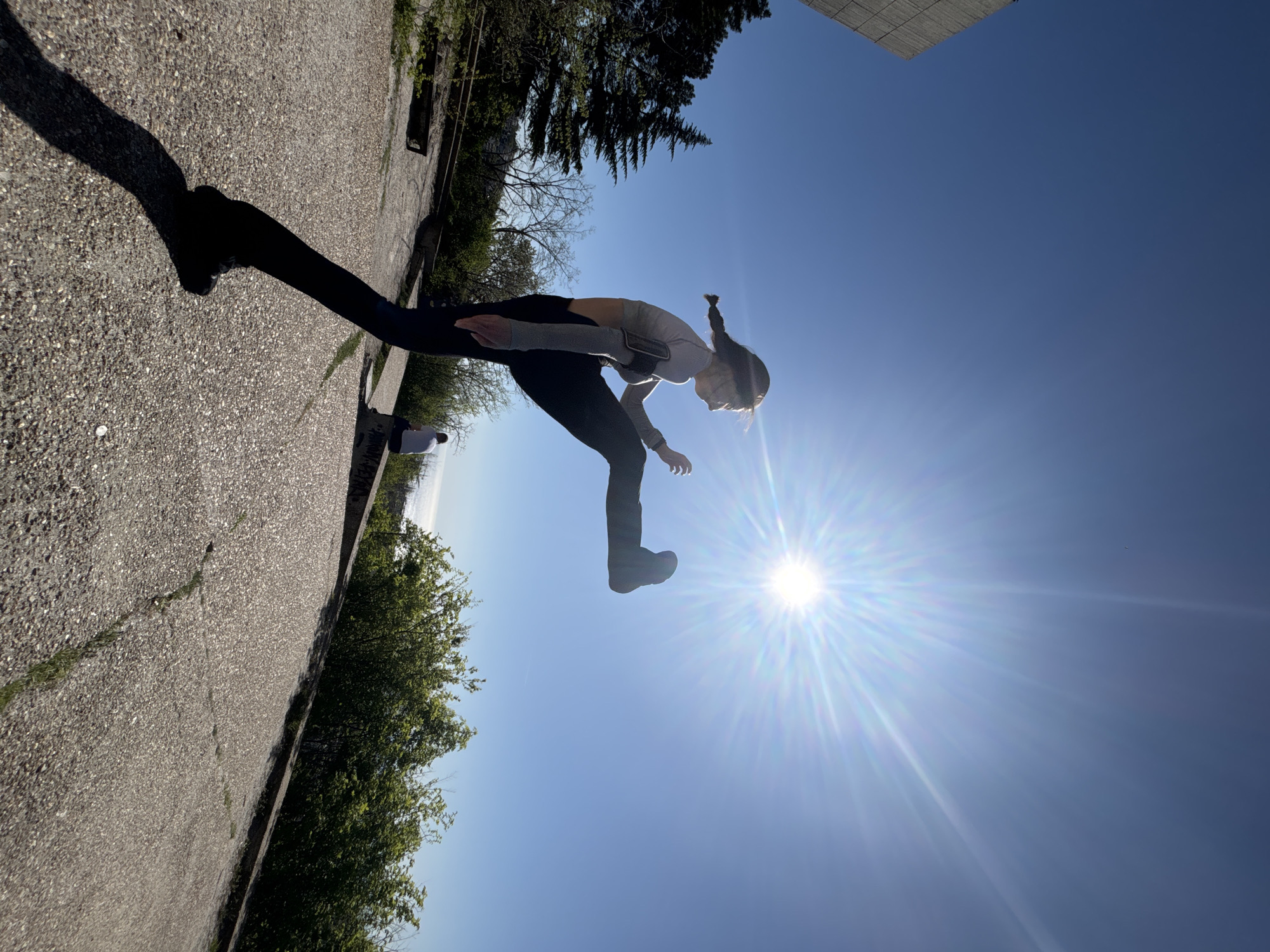 A young woman with her hair in pigtails is captured mid-jump against a clear blue sky, with the sun positioned directly behind her, causing a flare effect. She is wearing a long-sleeved shirt and dark pants. The ground appears to be a concrete surface, possibly a walkway or parking area, and it runs along the left side of the image. Trees and other greenery are visible along the edges, and a section of a building or structure is at the top right corner of the photo. The location is in the city of Unknown, Unknown. The image is taken from a low angle, emphasizing the height of the jump. The woman’s shadow is cast onto the ground to the left.