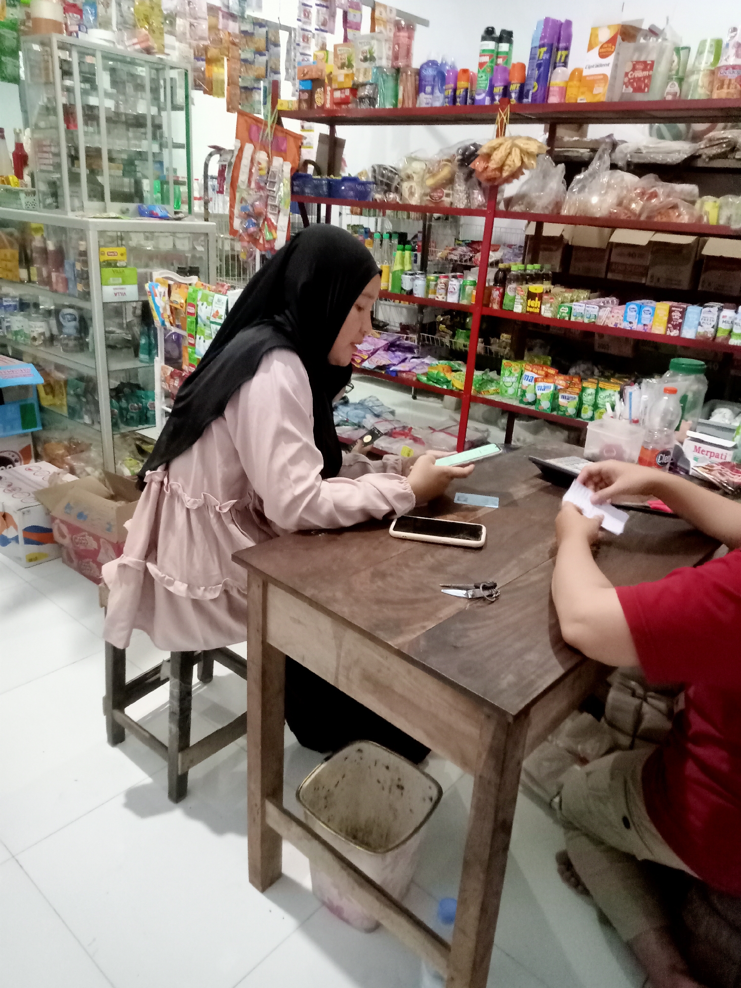 The image depicts an indoor scene, likely a small convenience store or warung, located in Simo, Indonesia. A woman wearing a black hijab and a pale pink, ruffled tunic is seated at a wooden table. She is holding a mobile phone in her right hand and appears to be interacting with it. On the table in front of her is a white smartphone placed face down and a pair of scissors. To her right, another person, whose upper body is visible in a red shirt, is holding a piece of white paper in their hands. Between the two individuals, a calculator is positioned on the table. The background reveals shelves stocked with various goods, including packaged food items, toiletries, and cleaning supplies, consistent with a retail environment. A plastic trash can is visible beneath the table on the left. The floor is tiled with white squares.