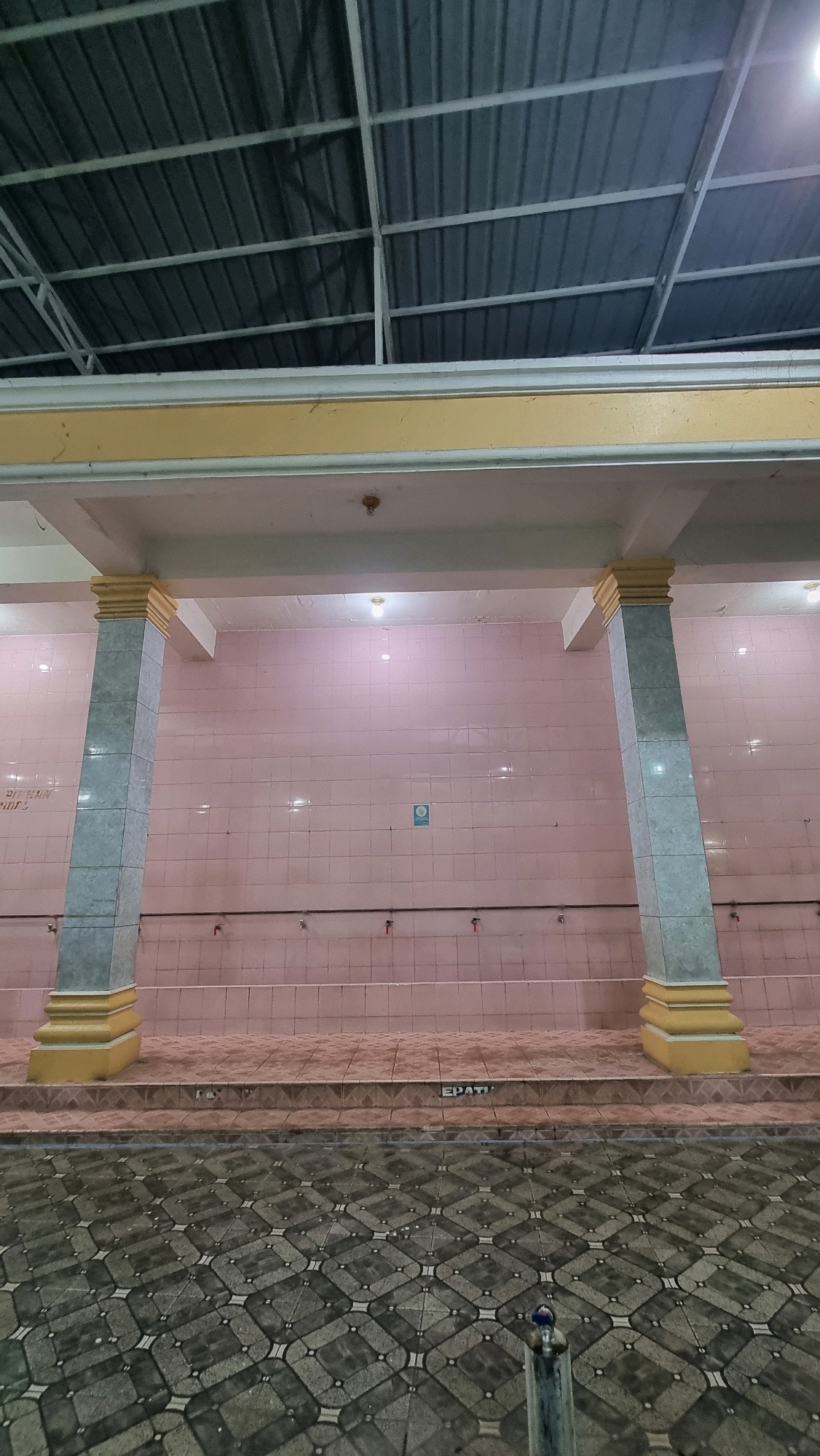 The image depicts an outdoor ablution area, commonly found in mosques or prayer facilities, in Puloara, Indonesia. The scene is characterized by a long wall covered in pale pink tiles, with a series of silver faucets mounted at intervals along its length. Two large, square pillars, clad in gray and white patterned tiles and topped with decorative yellow caps, stand prominently in the foreground, supporting a light-colored overhead structure. The floor in front of the tiled wall is also tiled, featuring a repeating geometric pattern in shades of gray and black. A step or platform leads down to a lower tiled area. The ceiling of the structure is visible, constructed from corrugated metal panels supported by a white metal frame. The lighting appears to be artificial, with visible light fixtures. The image is static and shows no people or discernible activity, focusing solely on the architectural elements and the clean, tiled environment.