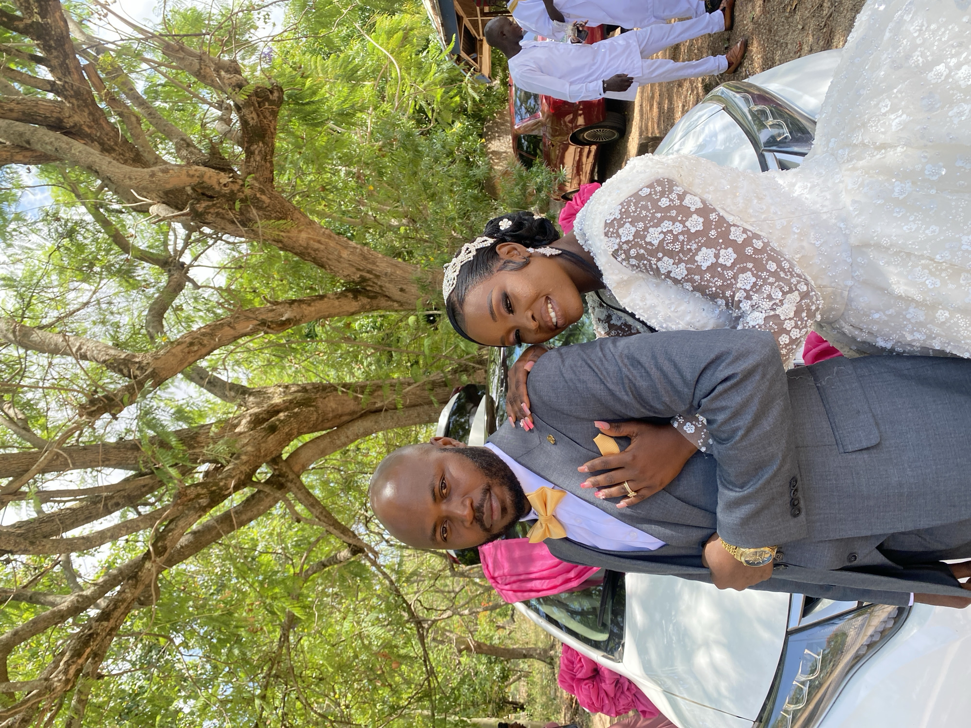 A wedding scene unfolds in Dutsen Kato, Nigeria. A couple, dressed in formal wedding attire, stands beside a white car adorned with pink fabric. The bride wears a white lace gown and a tiara, while the groom wears a grey suit with a gold bow tie and a gold watch. They are both smiling. Behind them are other people in white attire. A tree with expansive branches and green leaves provides a natural backdrop. A red car is also visible in the background.