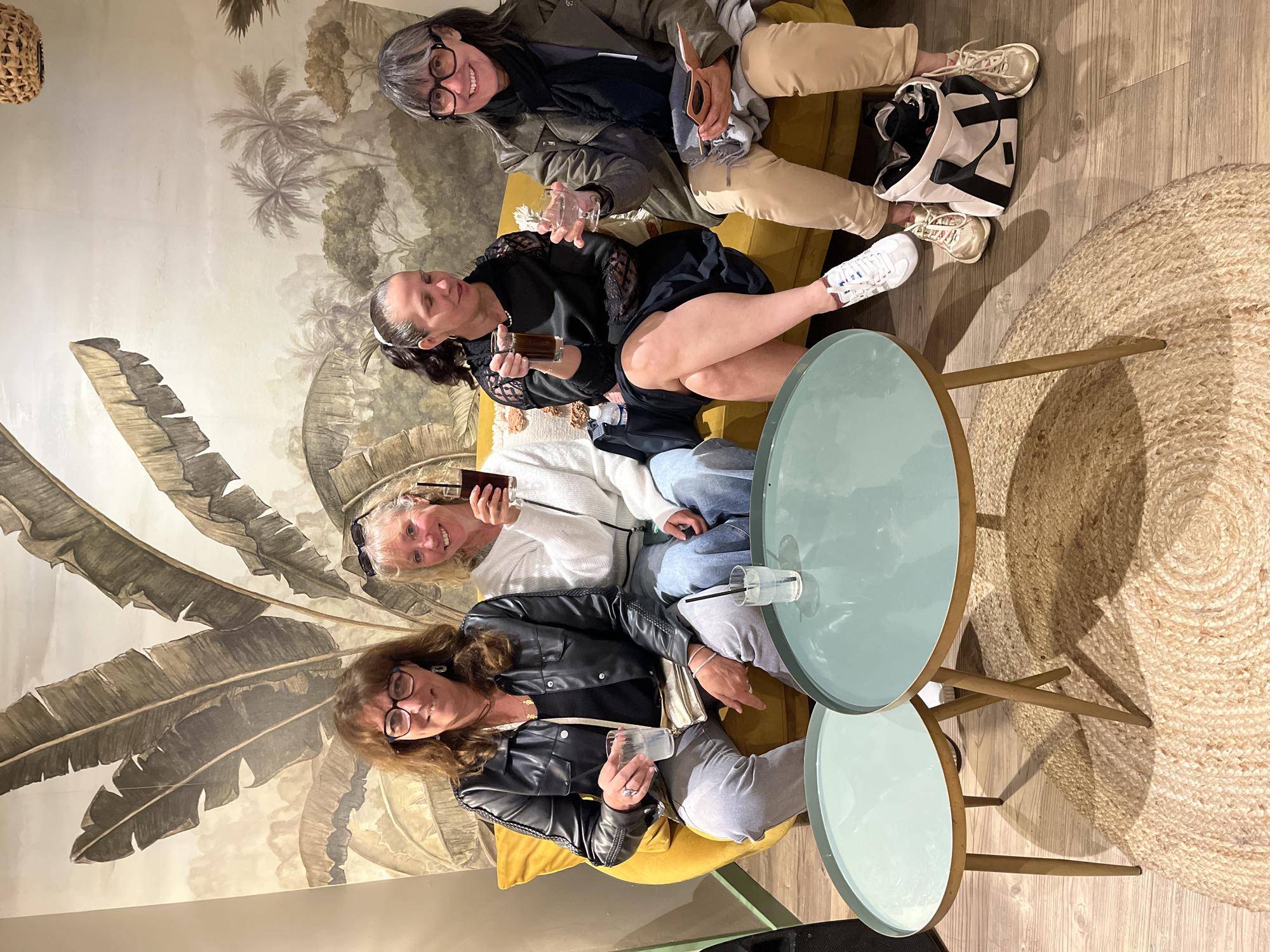 Four adult women are seated on a yellow sofa, possibly in an indoor establishment, based on the furniture and decor. They are holding drinks and appear to be socializing. The backdrop is a wall with a mural featuring tropical foliage, including banana leaves and palm trees. There are two tables with light blue tops nearby, and a round jute rug is visible. It is unclear whether this is Grimaud, France without further evidence.