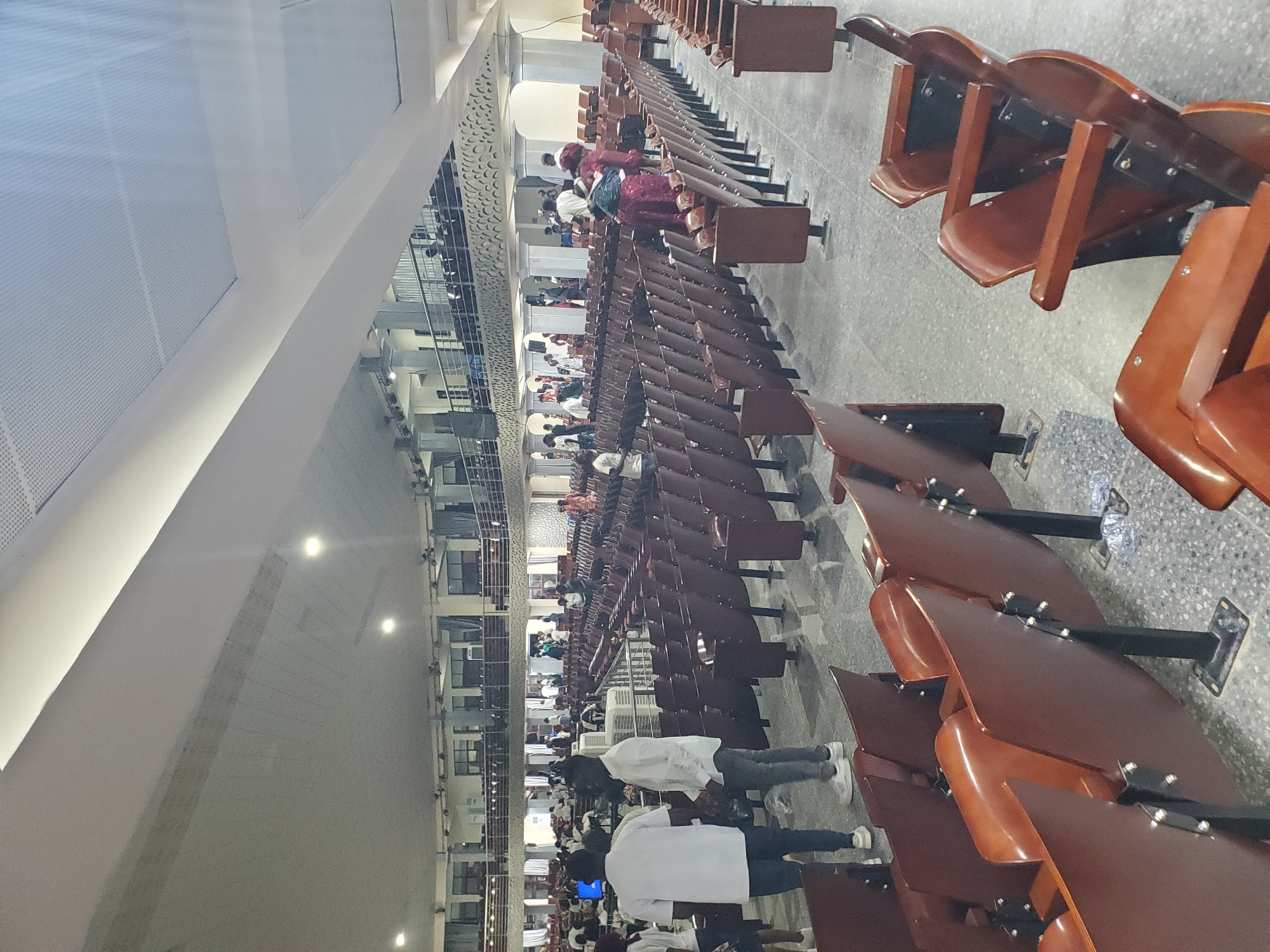 The image depicts the interior of a large hall, possibly a place of worship or a lecture hall, in Kason, Nigeria. The hall features rows of empty, dark brown wooden chairs with black metal legs, arranged in a semi-circular fashion. The floor is a speckled gray concrete. In the background, several people are visible, some standing and conversing, while others are seated on the upper level balcony. The lighting appears to be a mix of overhead fluorescent lights and some ambient light coming from windows or other openings. The architecture suggests a modern, functional space with a high ceiling and some decorative elements on the walls. The overall impression is one of an organized, possibly public gathering space.