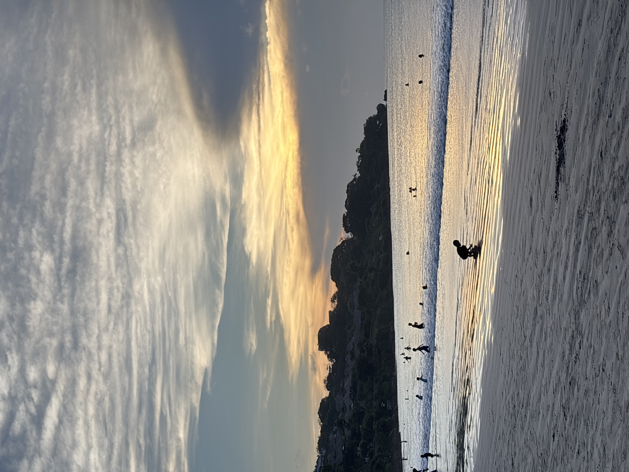 The image depicts a beach at sunset in Kuta Selatan, Indonesia. The sky shows a mix of white and golden clouds. A dark, tree-covered hill runs along the coastline, with buildings visible near the base. Several people are visible on the beach and in the water. The sand appears light-colored, and the water reflects the golden light of the setting sun. The image appears to be slightly rotated.