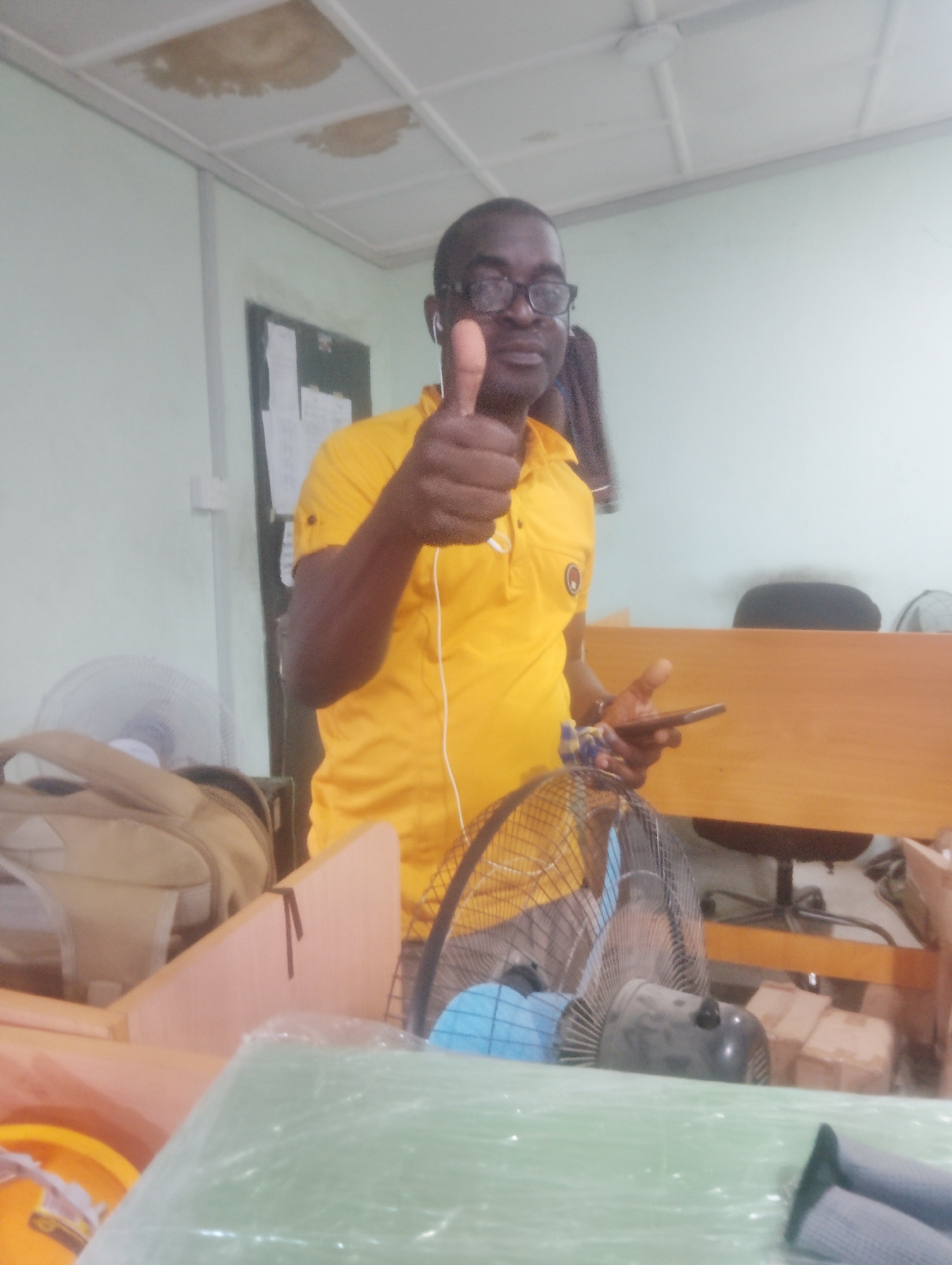 A medium shot captures a man in a yellow polo shirt wearing glasses and standing in what appears to be an office or storage space. He is holding a smartphone in his right hand and giving a thumbs-up with his left hand, suggesting a positive sentiment or approval. The background reveals a cluttered environment with office furniture, fans, bags, and cardboard boxes. The ceiling shows signs of water damage, with discolored patches indicating a potential leak. The scene is set in Ikeja, Nigeria, further suggesting it is an interior shot within a commercial or administrative building. The lighting is moderate, illuminating the man and his immediate surroundings, while the background remains somewhat dim.