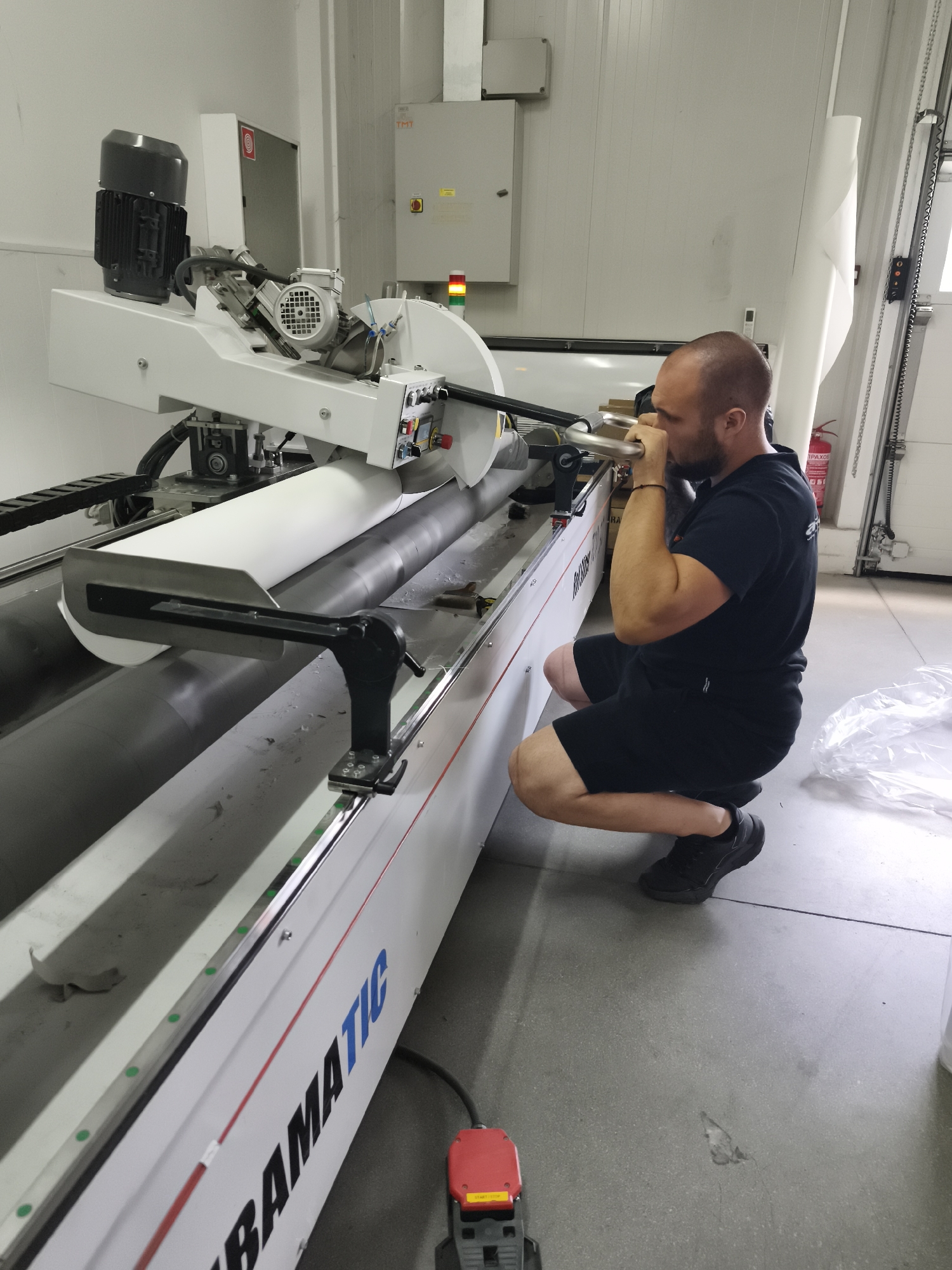 In an indoor industrial setting, a bald man with a beard, wearing a black t-shirt and black shorts, is crouching and operating a large white industrial machine. He is holding onto a metal bar with both hands and appears to be focused on his task. The machine has a large motor on top and a white roll of material on rollers. The floor is a light gray concrete. In the background, there is a white wall with an electrical panel and a fire extinguisher. The scene suggests a workshop or factory environment.