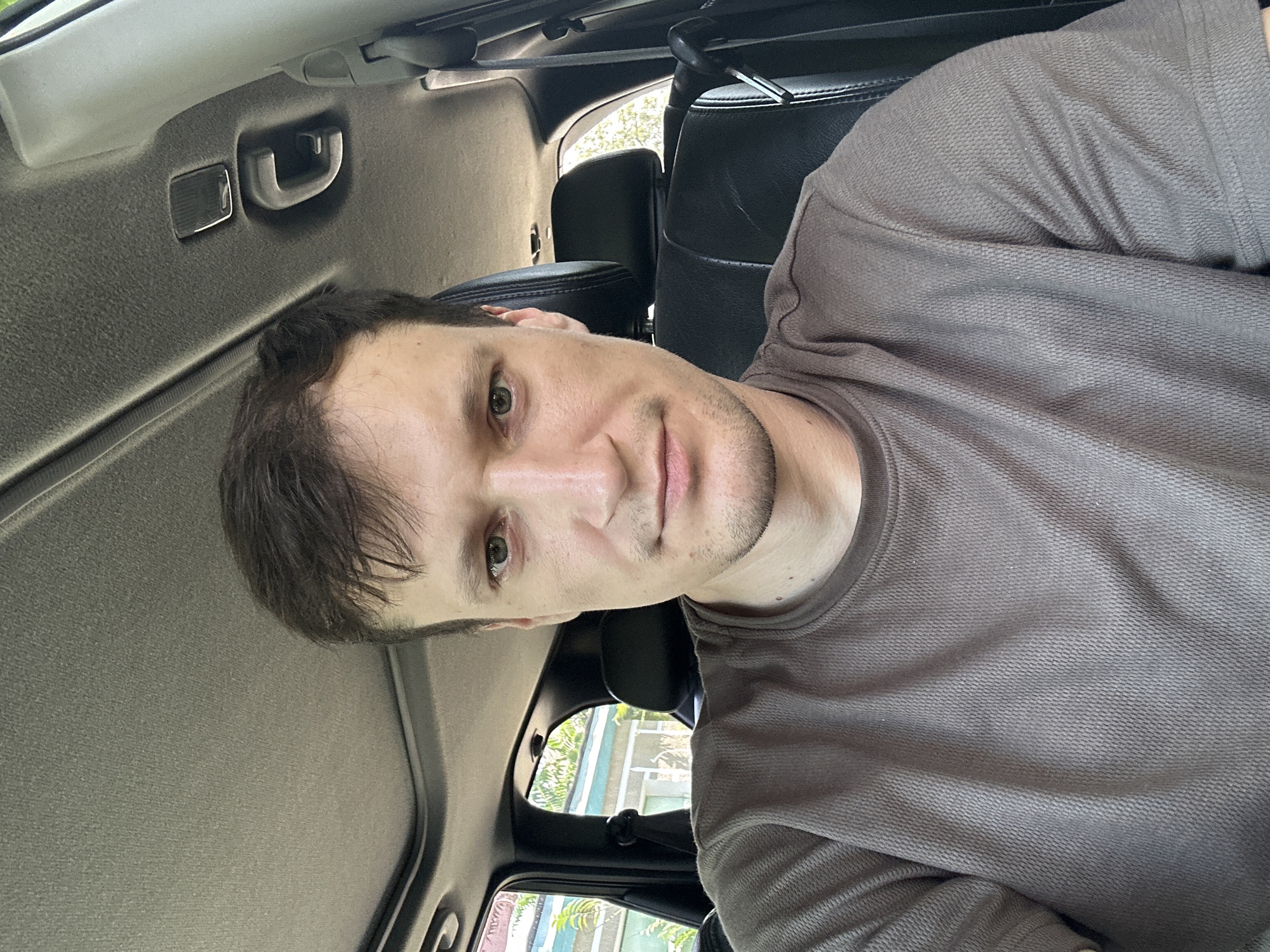 A close-up, forward-facing selfie shows a man with short, dark brown hair and green eyes sitting in the driver's seat of a vehicle. He is wearing a brown, textured t-shirt. The interior of the car is visible, including the sunroof, headliner, and black leather seats. The background outside the car shows greenery and parts of buildings, suggesting an urban or suburban setting. No specific location in Makiv, Ukraine, can be identified from the image.