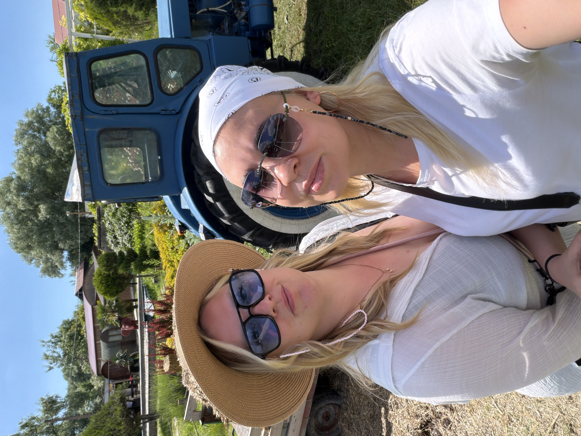 This outdoor photograph, taken in Gay-Kodzor, Russia, features two women posing in a sunny, rural setting.
On the left, a woman with blonde hair wears a white bandana adorned with small, repeating black motifs. She has on dark, rectangular sunglasses connected by a black and white beaded chain, a large gold hoop earring, and a white short-sleeved t-shirt. She looks towards the camera with a neutral expression.
To her right, another woman with blonde hair is wearing a wide-brimmed straw hat and dark, square-framed sunglasses, which are connected by a light pink beaded chain. She is dressed in a flowing, white, sheer-textured top or dress and also looks towards the camera.
In the left background, partially obscured by the women, is a blue tractor or similar agricultural vehicle, revealing its side and part of its cabin with multiple window panes. The background is filled with lush greenery, including various trees, shrubs (some evergreen, some with red leaves), and grassy ground. Further back, a building with a reddish roof, possibly a house or shed, is visible amongst the foliage. The bright blue sky and strong sunlight, casting clear shadows, suggest a midday or early afternoon setting. The overall scene depicts a relaxed outdoor moment. No discernible text is visible, though the bandana features decorative patterns.