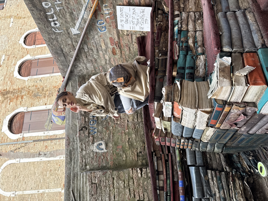 The scene depicts an outdoor alleyway within Libreria Acqua Alta in Venice, Italy. A young adult female is captured in a low squat, positioned on a stepped structure composed of numerous stacks of old, well-worn books of varying sizes and colors. Many book spines are visible, some appearing discolored or damaged. The top surfaces of these book stacks are partially covered with sections of worn, faded red fabric.
The female subject wears a beige, long-sleeved trench coat with a belt, layered over light blue distressed jeans featuring tears at both knees. A small, circular Louis Vuitton monogrammed shoulder bag with a tan leather tag is slung over her left shoulder, resting on her hip. Her left hand is raised, fingers touching her chin and neck area, while she looks directly forward. Her hair is styled in a bun, and she has visible makeup.
Behind her, a textured brick wall is present, displaying faint graffiti, including "WOU" in blue, and "GO UP". A diagonal metal pole or railing extends from the left midground upwards to the right, adjacent to further graffiti that includes "FOLLOW TH..." and an arrow pointing right. Below the railing on the right, a white rectangular sign with handwritten black text is affixed to the wall. It reads: "¡NO TE SIENTES, GRACIAS! DO NOT SIT DOWN. THANK U! NE T'ASSIEDS PAS. STP!"
In the upper background, a light brown brick building rises, featuring at least three evenly spaced arched windows with dark wooden shutters. A colorful piece of fabric, possibly laundry, is visible hanging near the center background. The scene is illuminated by natural daylight.