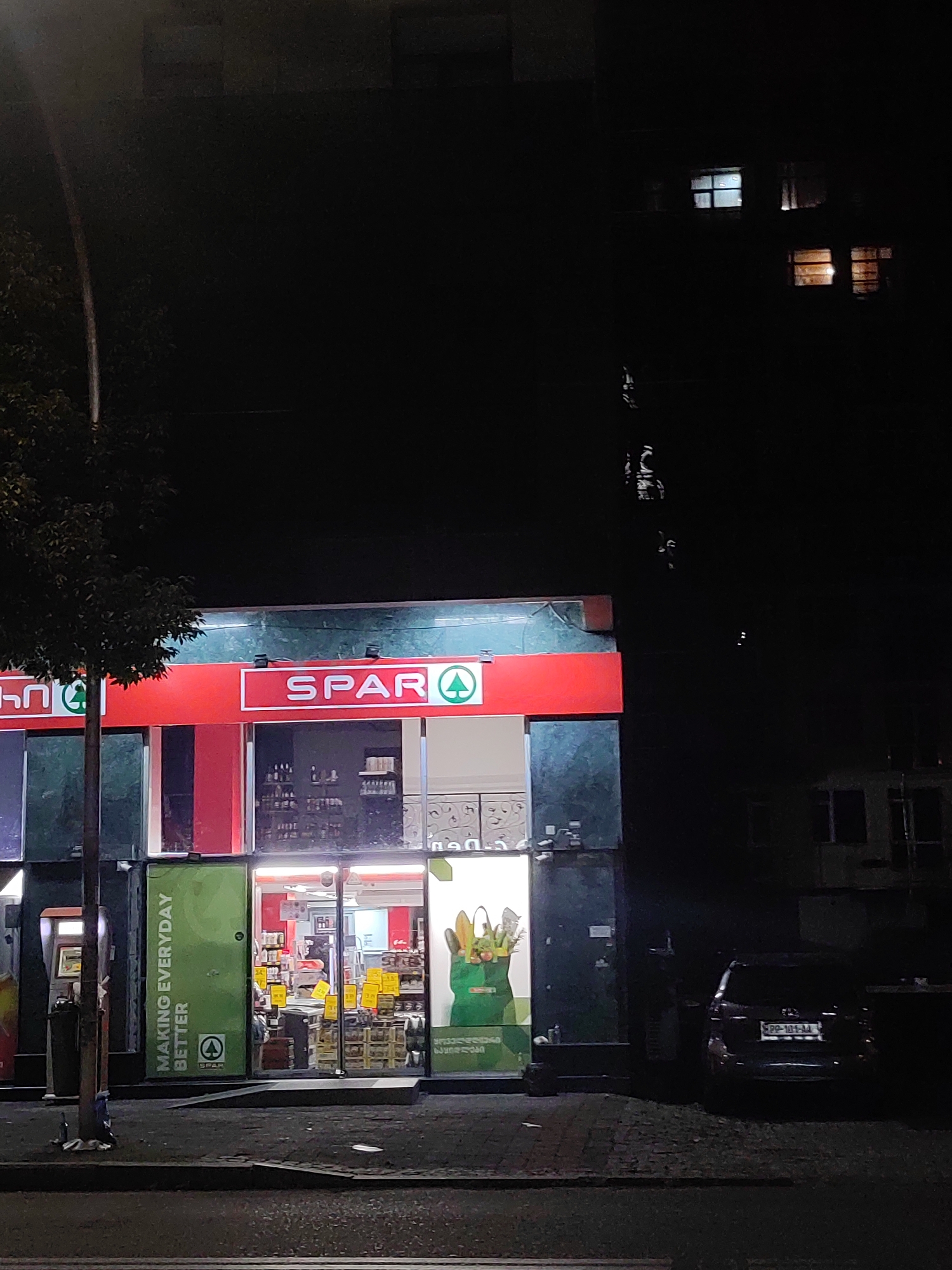 This is a nighttime outdoor, eye-level, medium shot of a SPAR supermarket in Batumi, Georgia. The store's red awning with white "SPAR" lettering is prominently displayed. Through the large glass storefront, shelves stocked with various items and several people can be seen. The SPAR logo, a green circle with a white outline, is visible on the awning and on green signage on a door to the left of the entrance. The green signage also has white text that reads "MAKING EVERYDAY BETTER". To the left of the store, an ATM machine is visible. A dark-colored car is parked to the right of the store. The street in front of the store is paved, and a tree with dark leaves stands to the left of the building. The background consists of a dark building with illuminated windows. The overall lighting suggests it is after dark.