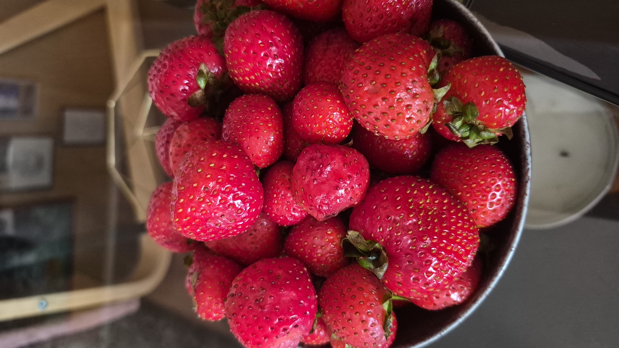 A close-up, vertical shot showcases a dark bowl overflowing with bright red strawberries. The strawberries vary in size and ripeness, with some exhibiting vibrant green stems and leaves. The focus is sharp on the fruit, with the background blurred and out of focus, suggesting a shallow depth of field. The lighting highlights the glossy texture of the strawberries, emphasizing their juicy appearance. The setting appears to be indoors, with hints of furniture and what might be a wooden object in the blurred background, suggesting a home or domestic environment. The location is Unknown, Unknown.