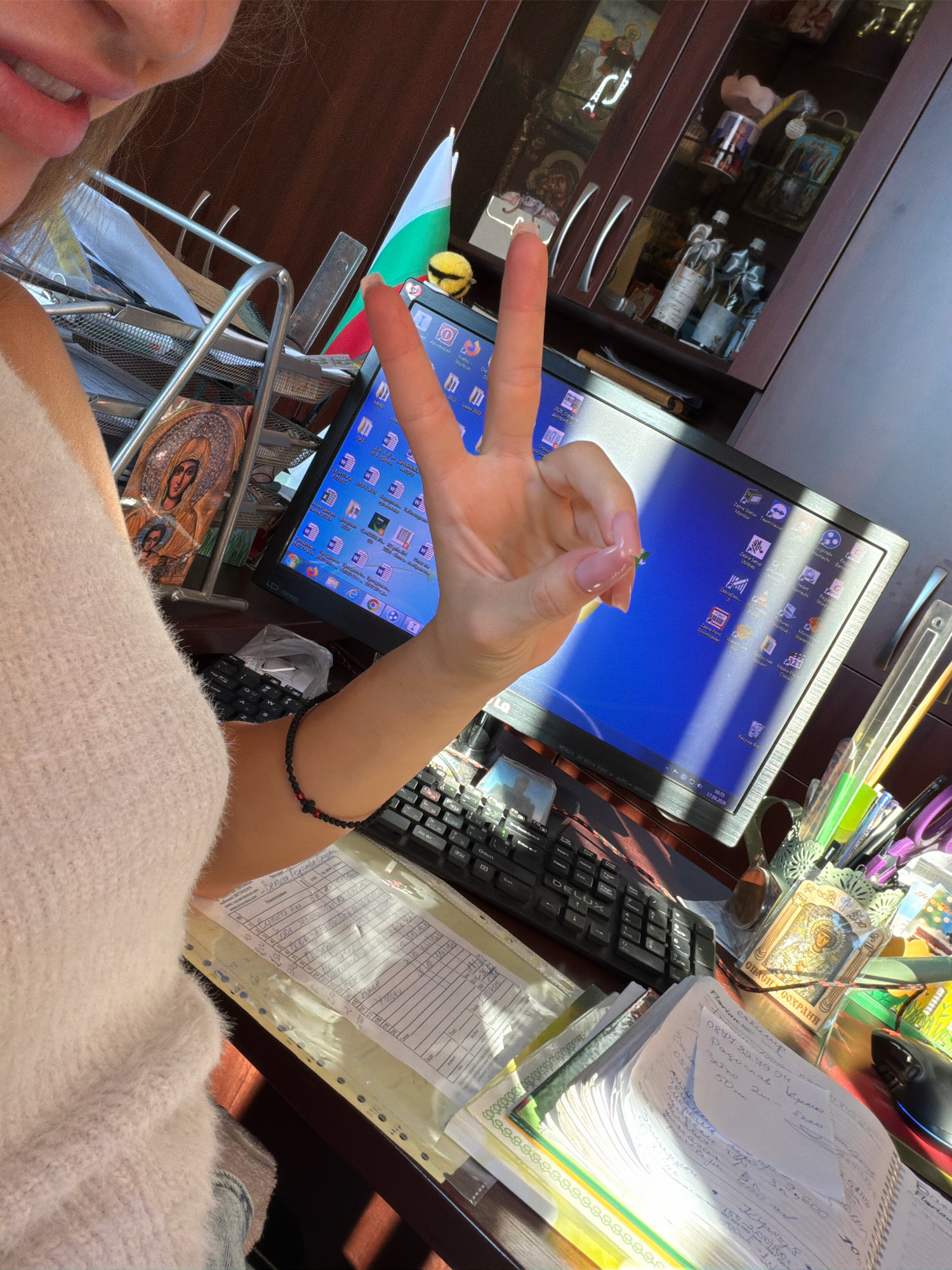 The image depicts a person's hand making a peace sign in front of a computer monitor, in what appears to be an office or home workspace. The workspace is cluttered with papers, a keyboard, pens, and other office supplies. A Bulgarian flag is visible in the background, suggesting the location is in Bulgaria. The computer screen displays a Windows XP-like desktop with several icons. The overall impression is one of a busy, personal workspace.