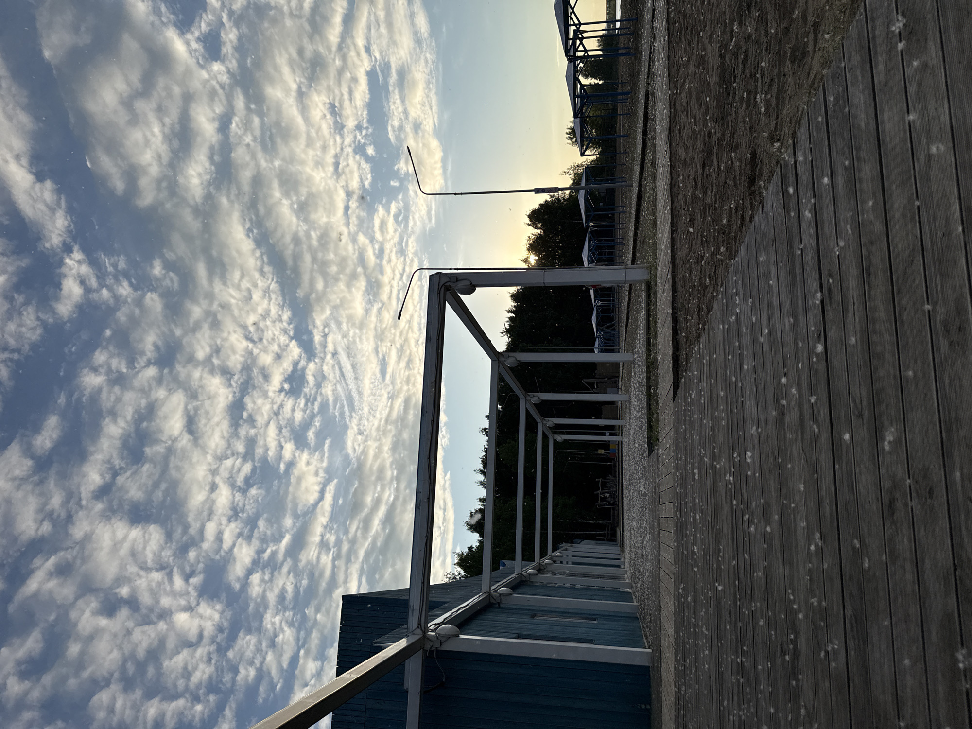 The image depicts an outdoor scene at a waterfront recreational area in the city of Chaikovsky, Russia. The time of day is either sunrise or sunset, evidenced by the low-angle, warm light on the horizon. The sky, occupying the upper portion of the frame, features a blue background largely covered by scattered white, textured altocumulus clouds.
In the immediate foreground, a dark wooden boardwalk composed of horizontally laid planks extends towards the mid-ground. To the right of the boardwalk is a dark brown sandy area. Both the boardwalk and the sand are sparsely covered with numerous small, white, fuzzy particles, consistent with botanical fluff (e.g., poplar seeds).
On the left side of the image, a building with vertical blue paneling is partially visible. Attached to and extending from this building is a light-colored metal pergola structure, characterized by a series of rectangular frames supported by vertical posts. Electrical wires are visibly strung along the top beams of this structure.
In the mid-ground, two tall, dark-colored streetlights with curved arms and lamp heads are prominently silhouetted against the sky. Further to the right and in the background, a series of identical open-sided shelters with blue frameworks, likely beach canopies, are arranged on the sandy terrain. A dense line of dark green trees forms the backdrop, through which the low sun is partially visible on the right, casting an orange glow on the distant horizon. A body of water is faintly discernible in the far right background, beyond the trees and shelters. No individuals are present within the frame.