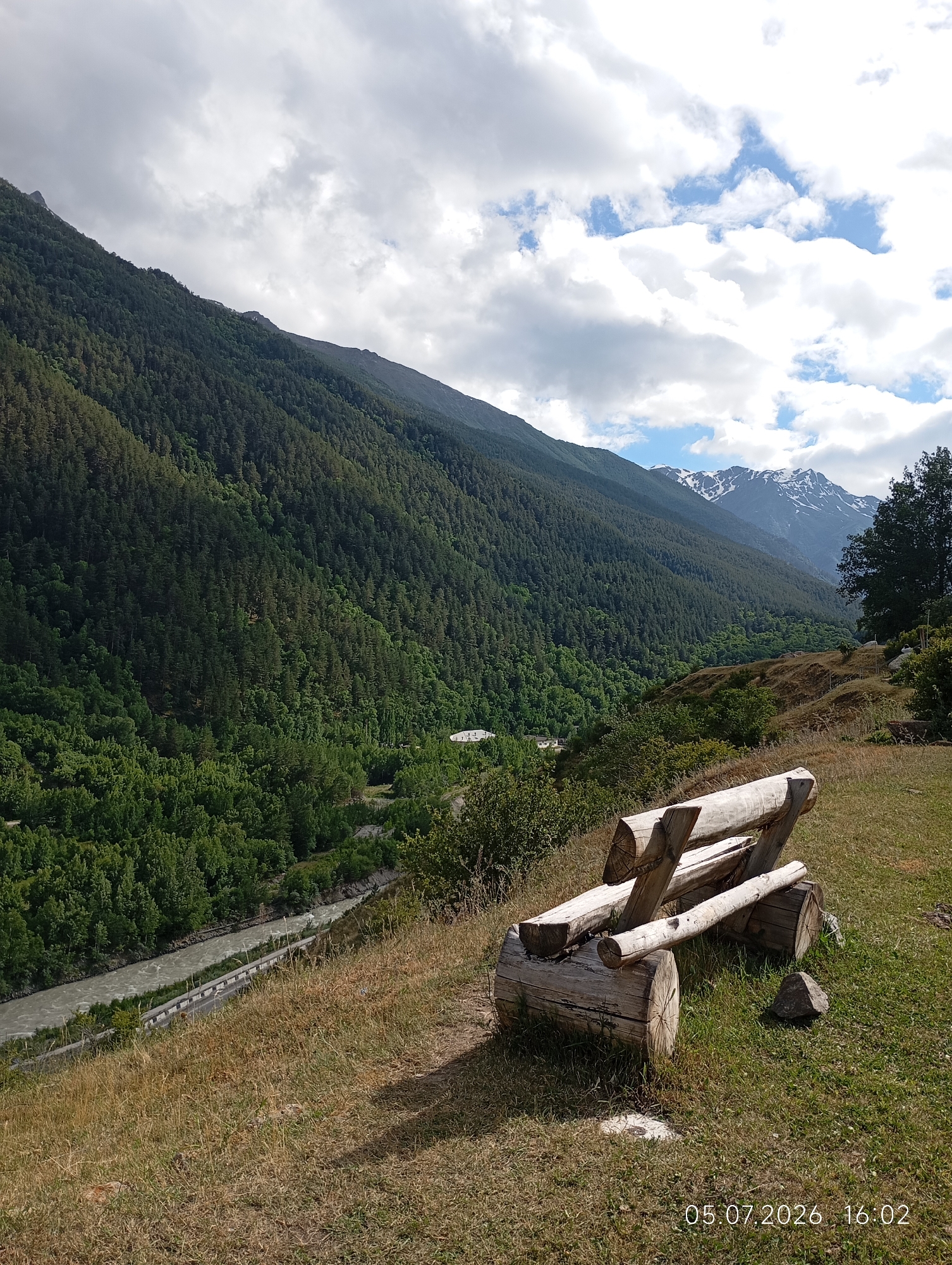In the city of Neutrino, Russia, an outdoor scene is captured. In the foreground, a rustic wooden bench, constructed from logs, sits on a grassy slope. The bench is positioned facing away from the viewer, towards a vast mountainous landscape. The mountains are covered in dense green forest, with a winding river visible in the valley below. The sky above is partly cloudy, with patches of blue sky interspersed with white clouds. In the distance, snow-capped mountain peaks are visible. The overall atmosphere is serene and picturesque, highlighting the natural beauty of the region. The date and time stamp "05.07.2026 16:02" is present in the bottom right corner of the image.