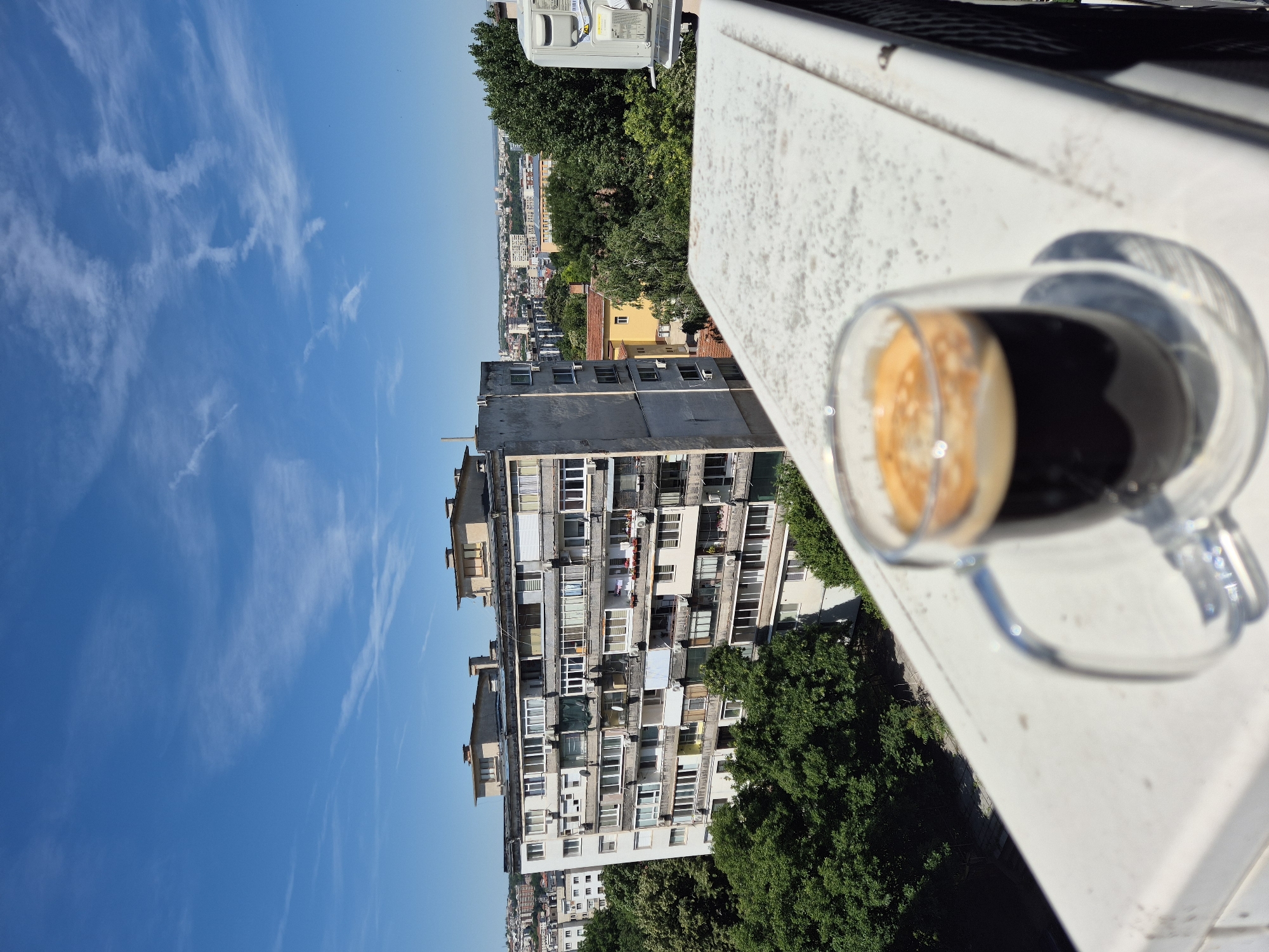 An eye-level, full shot depicts a clear, bright day with a city skyline in the background and a glass of espresso in the foreground. The espresso cup, made of clear glass with a handle, sits on a weathered white ledge. The dark espresso has a frothy, light brown crema on top. The city skyline features a large, multi-story concrete building with numerous windows, balconies, and air conditioning units. Dense green trees are scattered throughout the city, and a few smaller buildings are visible in the distance. The sky is a vibrant blue with thin, wispy white clouds. The ledge in the foreground is textured with a white, speckled surface and has a dark railing on the right side.