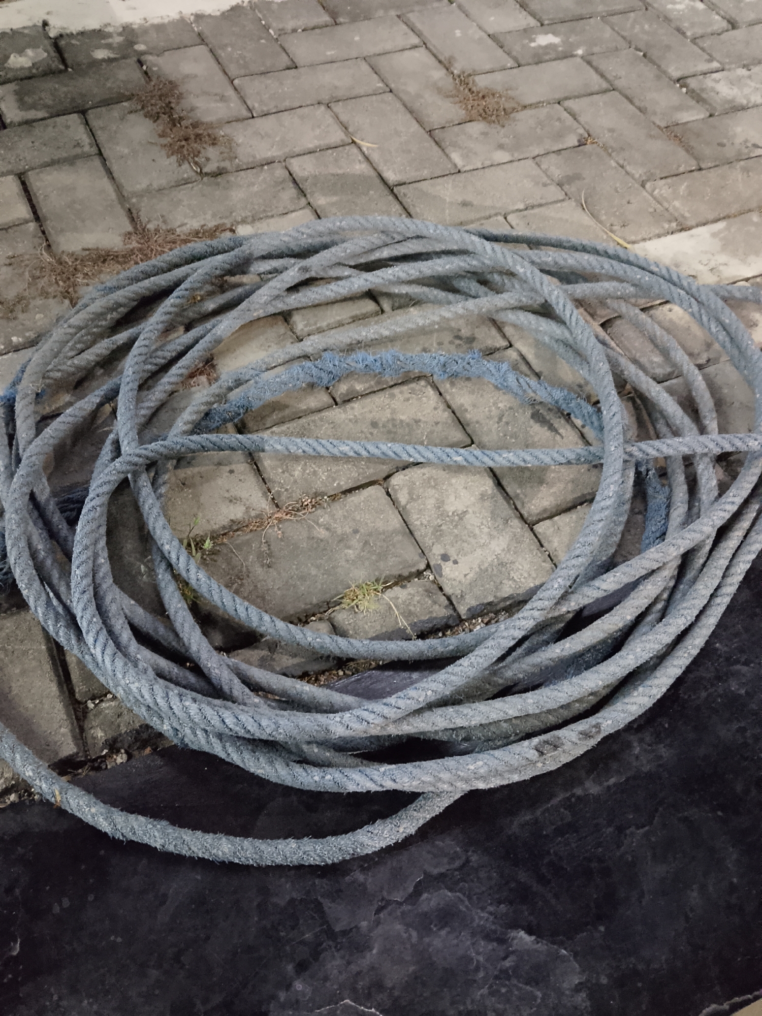 A coil of thick, grey rope is laid out on a paved surface in Kasiman, Indonesia. The rope is circular and appears to be made of synthetic fibers, with some fraying visible. The paved surface consists of rectangular interlocking stones, some of which are covered with a thin layer of dry leaves and debris. To the right of the paved area, a dark, flat surface is visible, possibly asphalt or a tarpaulin. The scene is outdoors, and the lighting suggests it may be evening or nighttime, with some artificial light illuminating the area. No people or animals are present in the image.
