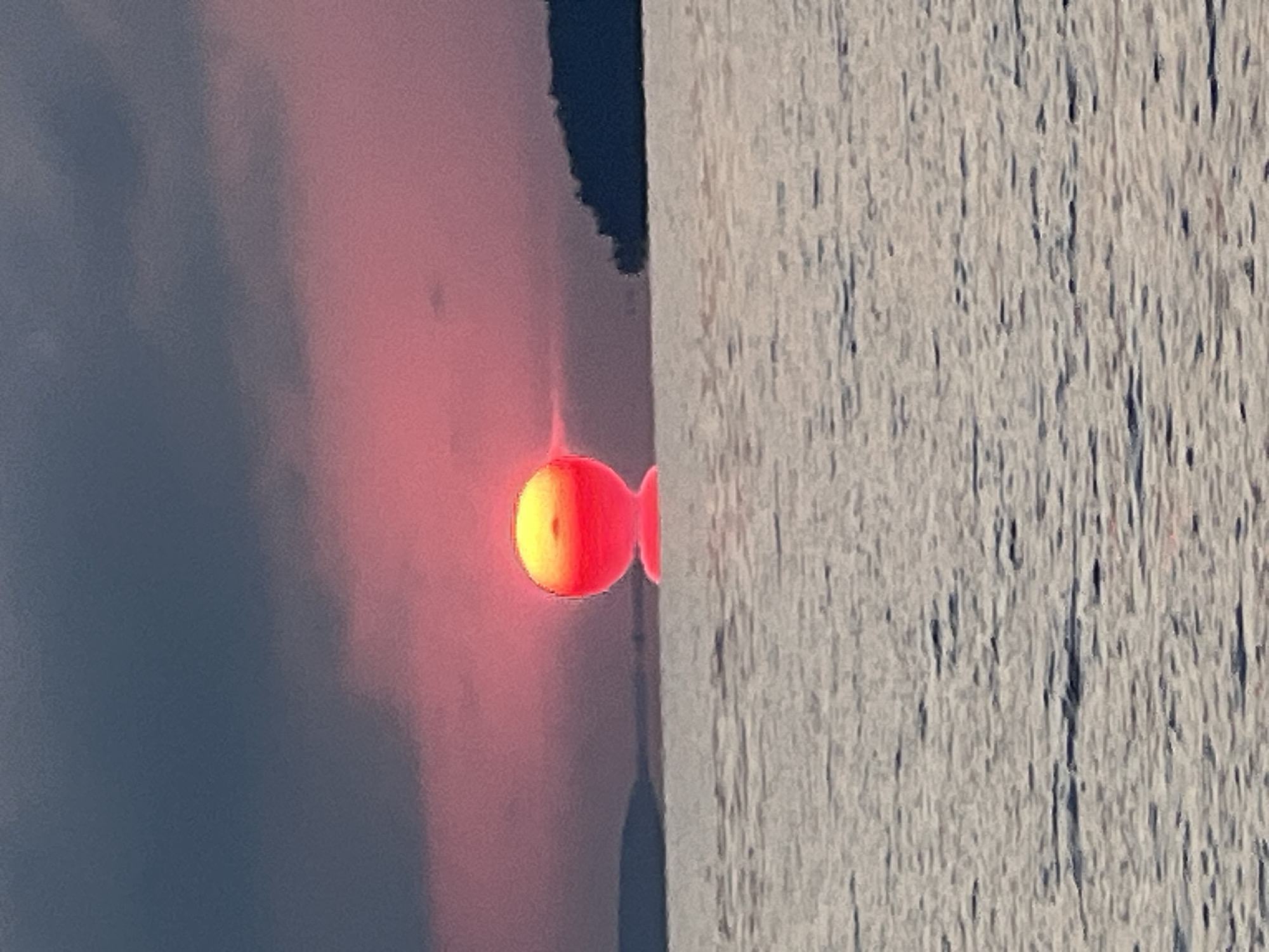 A sunset is seen over a body of water, likely a lake or sea, near the city of Novopichugovo, Russia. The sky is a gradient of dark blue and pink, with clouds obscuring the upper portion. The sun, a vibrant orb of yellow and red, is positioned just above the horizon, casting a pink glow. A faint reflection of the sun is visible on the water's surface. In the distance, dark silhouettes of landmasses are visible on either side of the sun. The water itself is a choppy expanse of grey and white, reflecting the dim light of the setting sun. No people or man-made structures are visible in the frame.