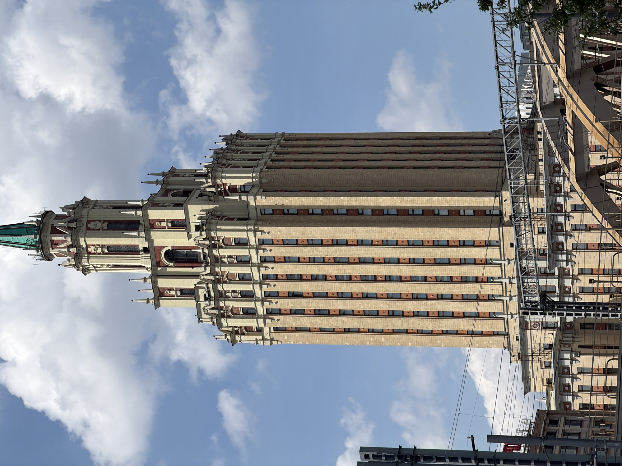 The image shows the Kotelnicheskaya Embankment Building, one of the Seven Sisters skyscrapers in Moscow, Russia, against a backdrop of blue sky with scattered clouds. The building is viewed from a low angle, emphasizing its height and architectural details. It features a light-colored facade with repetitive window patterns and decorative elements, including spires and ornate detailing near the top. Scaffolding is visible on one side, indicating ongoing maintenance or construction work. Other buildings and structures are visible at the bottom of the frame. The image captures a clear, daytime scene.