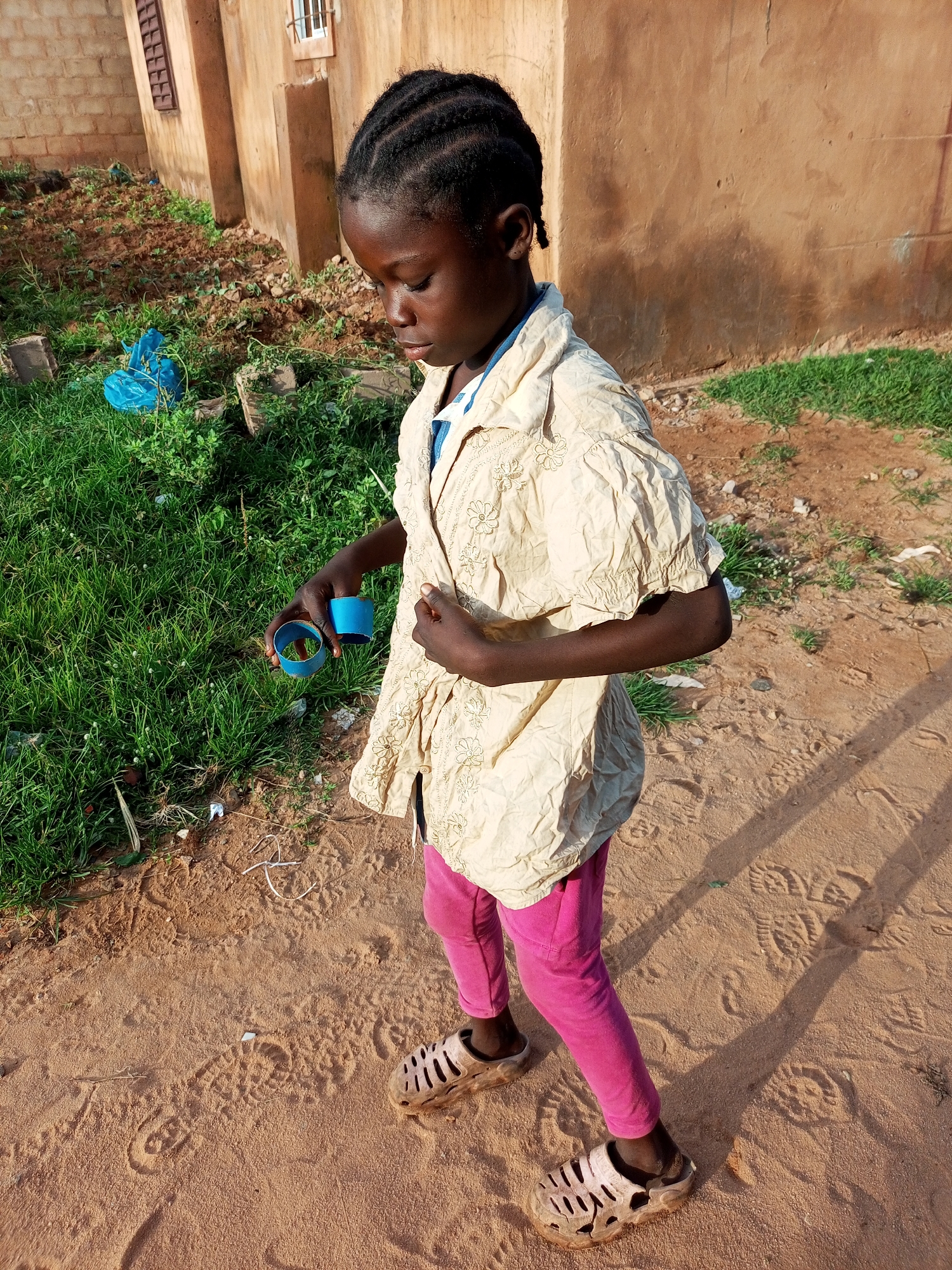 A young Black girl, with braided hair, stands in a sandy outdoor area in front of a tan-colored building. She is wearing a cream-colored, patterned shirt and pink leggings. Her feet are clad in light-colored, perforated sandals. The girl is holding two blue cylindrical objects in her right hand. The ground is sandy with visible footprints and some patches of green grass and dirt. Shadows of the building and possibly the girl are cast across the sand. The building has a dark-slatted window. A blue plastic bag is visible on the grass to the left. The image appears to be taken in Tempure, Nigeria.