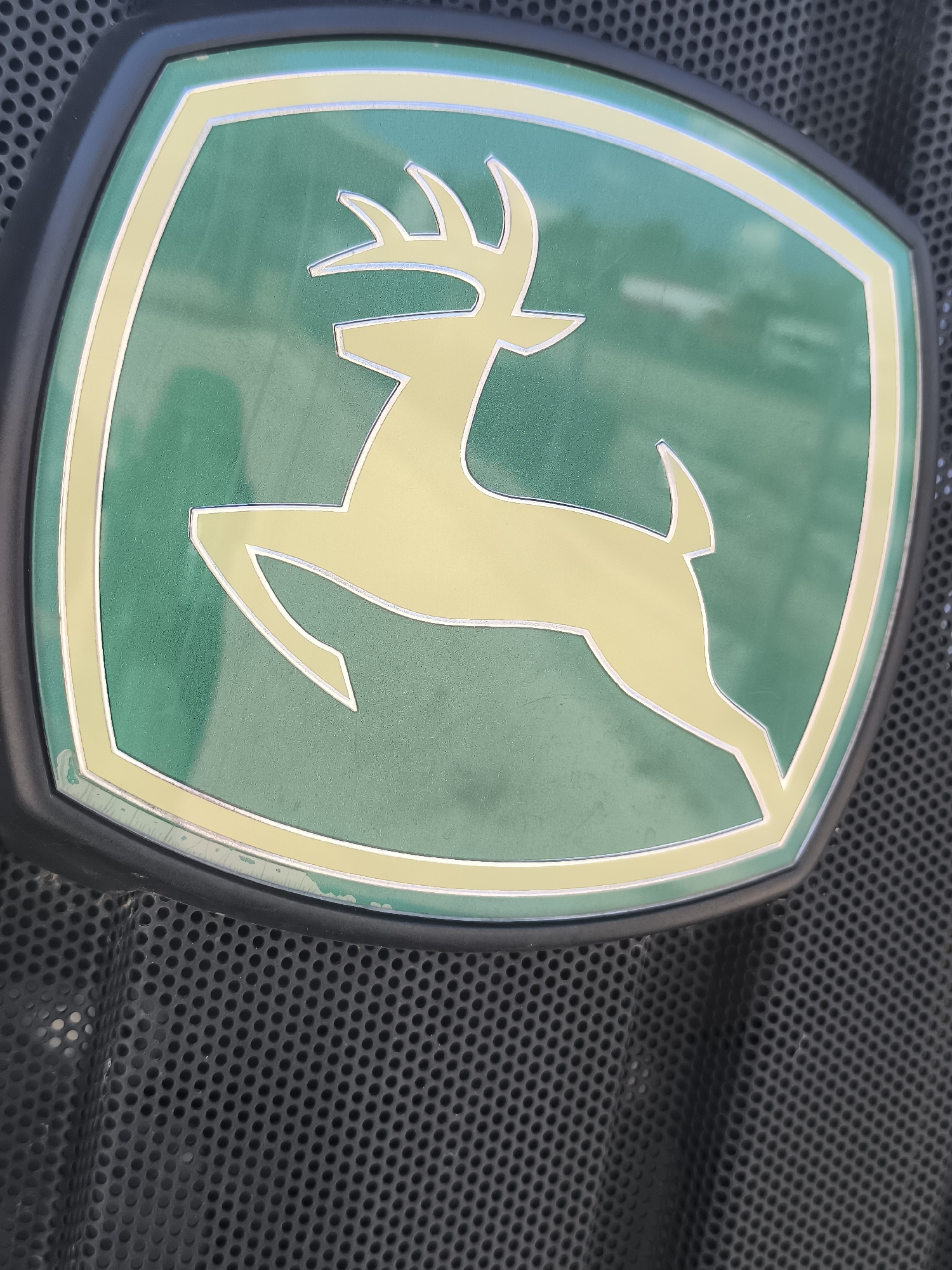 The image is a close-up, angled shot of a yellow deer silhouette on a green background, enclosed within a golden border. This emblem is bordered by a black frame and is affixed to a black perforated surface, which appears to be a grill or housing. The emblem itself exhibits some minor wear and tear along the bottom edge. The background is blurred, suggesting it is outdoors, but no specific urban features or landmarks are visible that would allow for a precise location within Maior Uzunovo, Bulgaria. The scene is static and focuses solely on the emblem and its immediate surroundings.