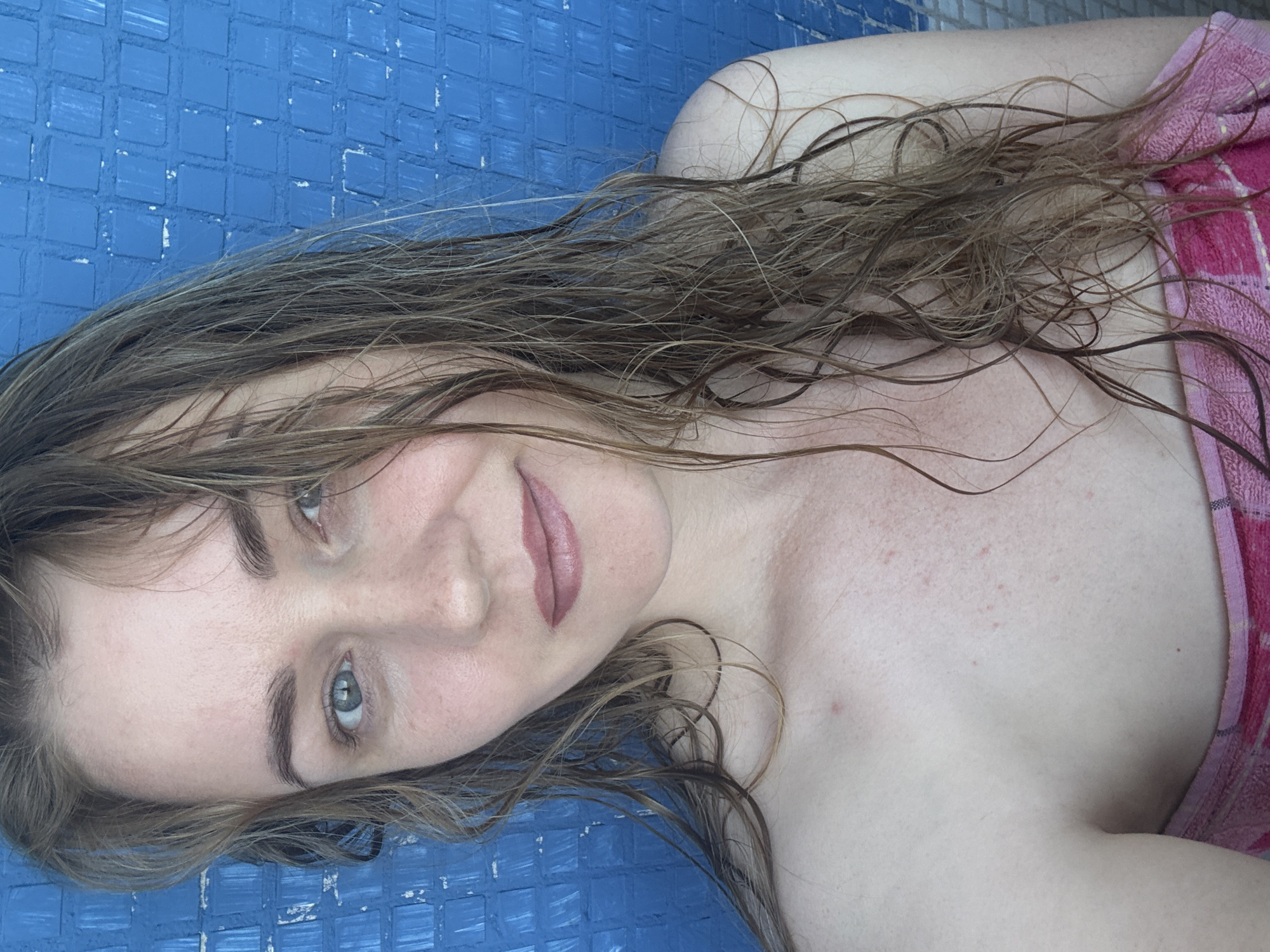 A woman with wet, brown hair is shown from the chest up. She is looking at the camera with a neutral expression. She is wearing a pink towel draped around her shoulders. The background is a blue tiled wall. The image is well-lit and in focus.