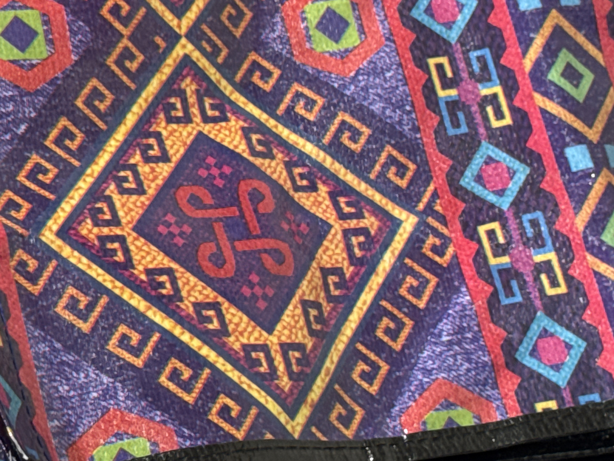 The image is a close-up, overhead shot of a fabric with a vibrant, geometric pattern. The pattern features a central square containing a red, interlocked symbol on an orange, textured background, surrounded by a yellow border. This central motif is framed by a larger purple area with repeated, angular yellow Greek key-like designs. Diagonal lines and triangular shapes in red and orange also break up the pattern. Toward the bottom of the image, a band of colorful geometric shapes, including diamonds and abstract zigzags in red, blue, pink, and green, is visible. The fabric appears to be textured, with a slight speckling effect on the purple areas. The lighting suggests it is indoors or in a shaded outdoor area. There are no people or specific landmarks identifiable to place it within Tbilisi, Georgia. The image focuses solely on the textile design.