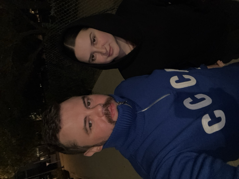 Two people pose for a selfie at night. The man is wearing a blue Gucci turtleneck sweater with a zipper. The woman is wearing a black hoodie. The photo is taken at an angle, with the subjects slightly rotated. The background is dark, possibly a street or park.