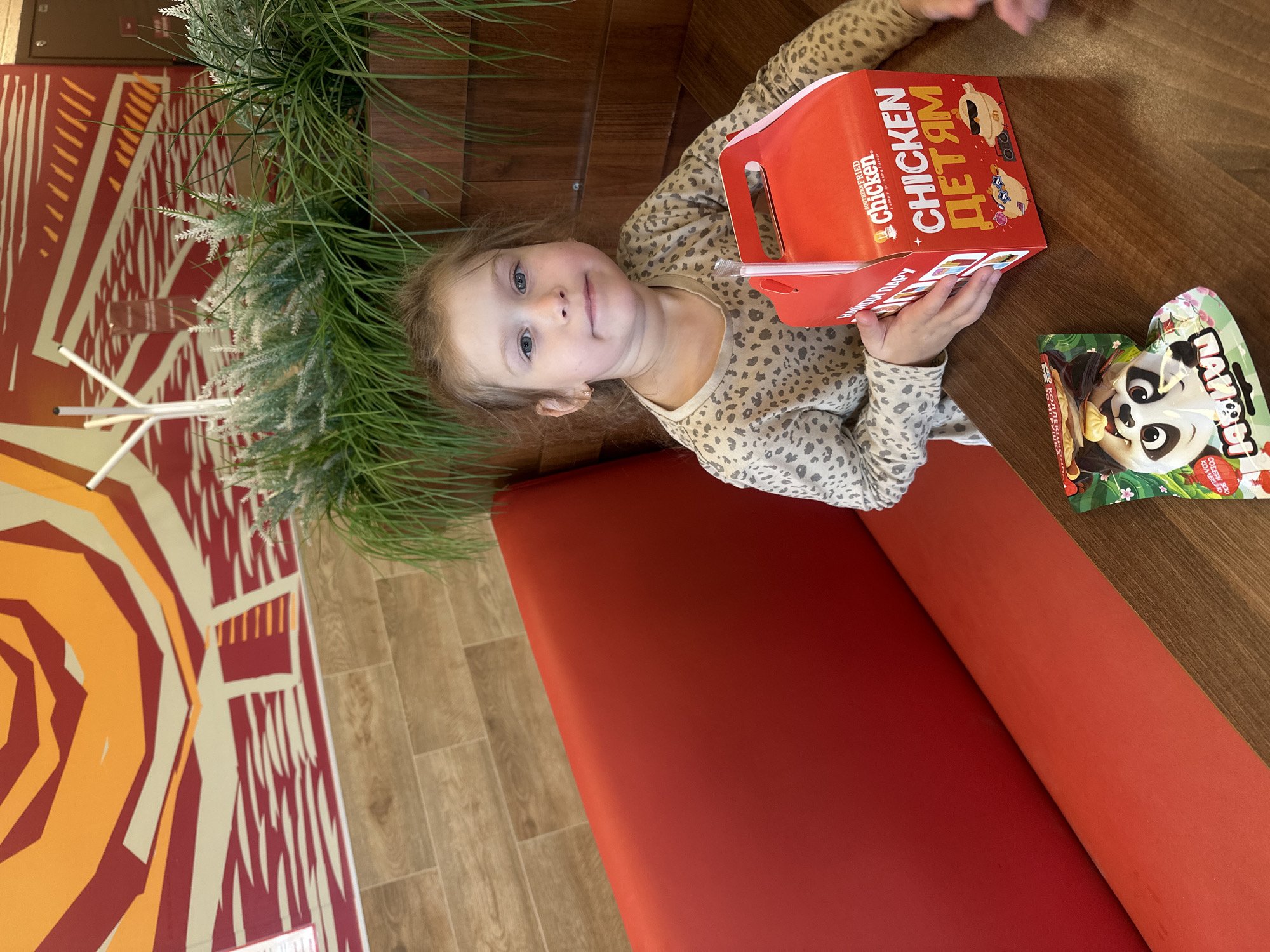 A young girl with light brown hair and blue eyes sits at a wooden table in a restaurant booth. She is wearing a tan and grey leopard-print long-sleeved shirt. In front of her is a red cardboard box with a straw sticking out of it. The box has the words "SOUTHERN FRIED Chicken" and "CHICKEN ДЕТЯМ" printed on it in yellow and white lettering, along with cartoon images of chickens. To the left of the box is a small, colorful snack packet featuring a cartoon panda. The girl is looking directly at the camera with a neutral expression. The background shows a red booth seat, wooden paneling, and a large, abstract mural with orange and red geometric shapes on a cream background. Artificial green plants are visible behind the girl. The scene is set within the city of Krilovo, Russia.