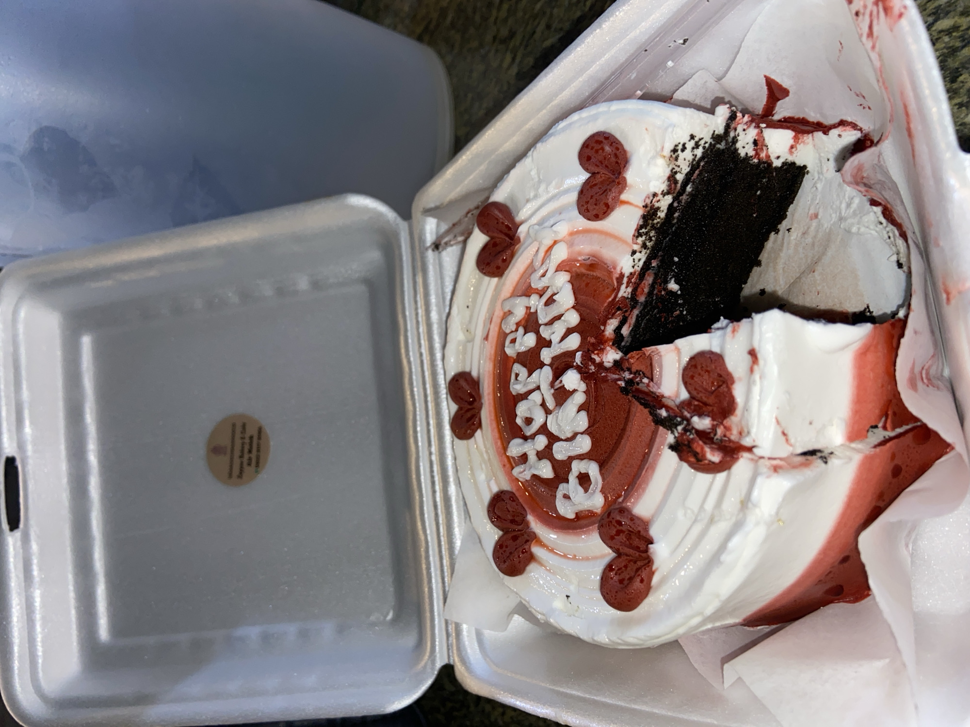 In the city of Candirejo, Indonesia, an opened white styrofoam take-out container sits on a surface. Inside the container is a partially eaten round cake with white frosting and red accents. The cake has "Happy Birthday" written on it in white frosting. There are several red heart-shaped decorations on top of the cake. A slice has been removed from the cake, revealing a dark chocolate interior. The styrofoam container has some red frosting and cake debris on its edges and inside. A light blue plastic jug is visible in the background, partially obscured by the open lid of the styrofoam container. A small circular sticker with text is adhered to the inside of the lid.