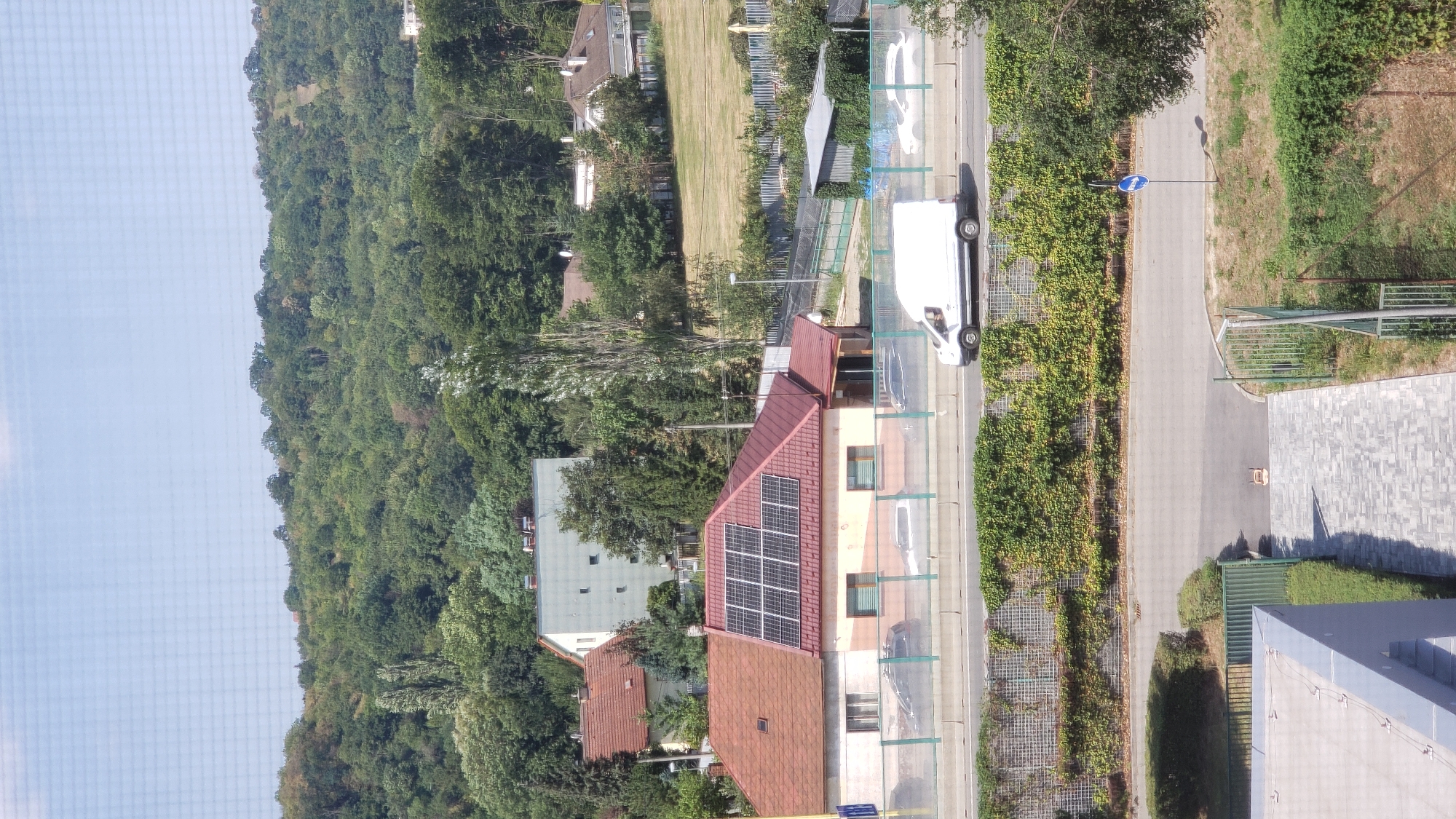 The image shows a residential area with a hilly, tree-covered background. In the foreground, there is a multi-story building with a gray facade and a paved walkway leading to it. Adjacent to this building is a street with a green fence and a wall covered in ivy. Beyond the fence, a white van is driving on a road. Further back, there is a house with a red roof covered in solar panels, and several other houses and buildings are visible among the trees. The sky is clear and blue. This scene is not in Prešov, Slovakia, as no specific landmarks or information indicate this location.