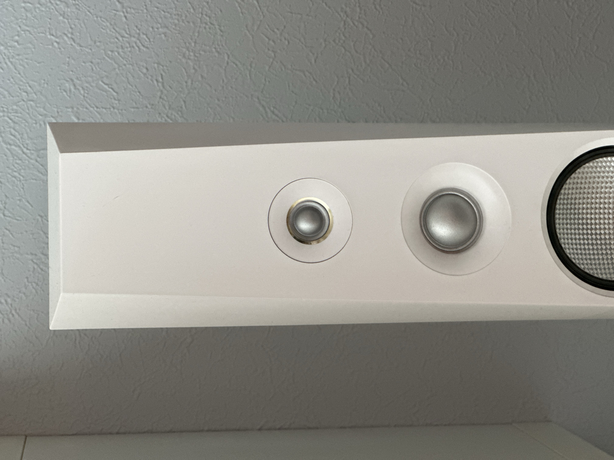 A vertical, close-up, eye-level shot shows the front of a tall, white speaker. The speaker has a tapered shape, wider at the bottom. Two circular speaker drivers are visible, each with a silver cone. The top driver is surrounded by a thin gold ring. Below the second driver, a portion of a third driver is visible, featuring a textured grille. The speaker is positioned against a textured, light gray wall. No people or animals are present in the image.