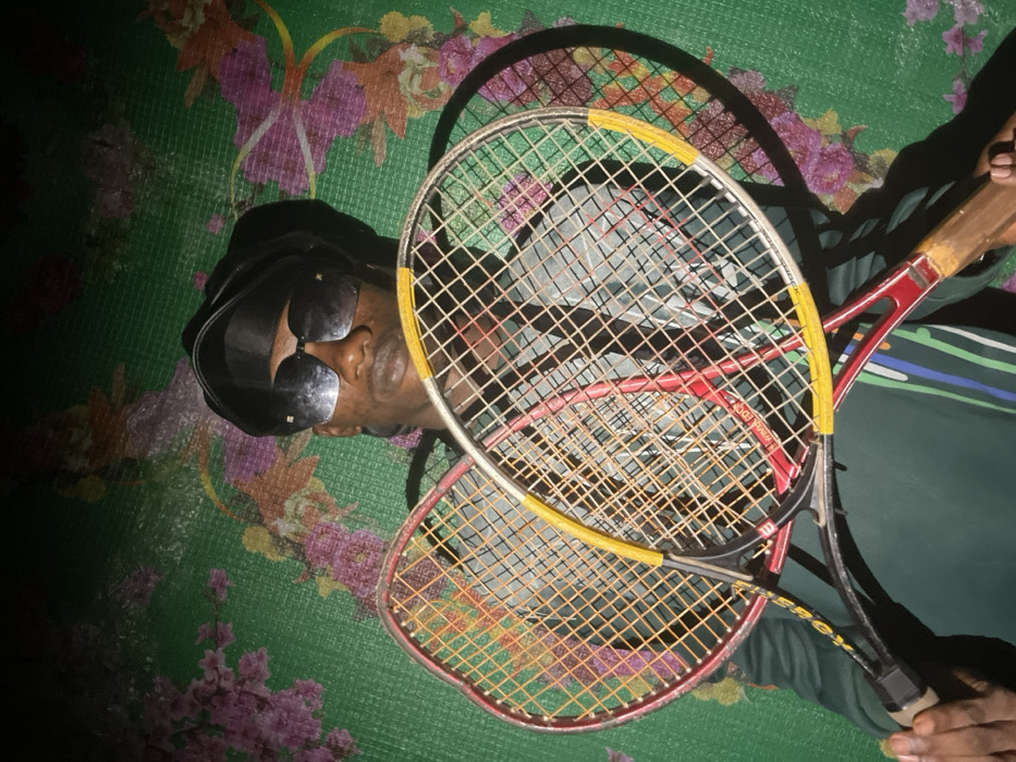 An overhead shot displays a person in sunglasses and a dark head covering, lying on a green floral-patterned surface. Two tennis rackets overlap, partially obscuring the person. The top racket has a yellow frame, while the one underneath is red. Shadows from the rackets are visible on the surface. The person's hand holds one of the racket handles. The light source is angled, creating contrasting light and dark areas.