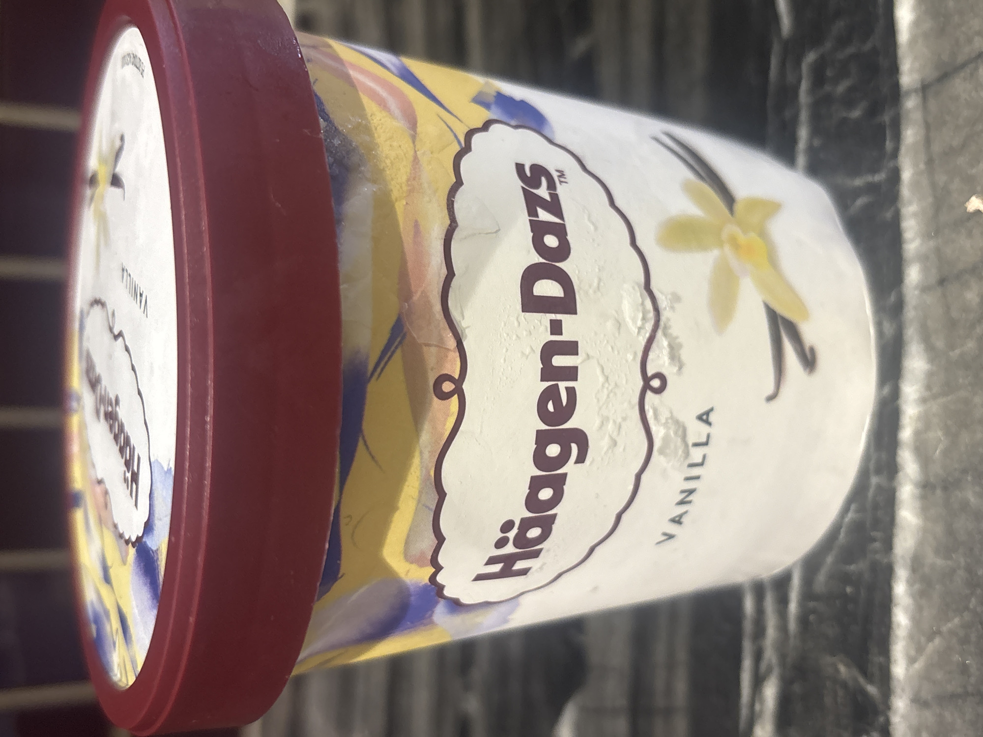 The image is a close-up, side-view shot of a pint of Häagen-Dazs vanilla ice cream. The container is mostly white with a red lid. The label features the Häagen-Dazs logo in a dark purple script, with the word "Vanilla" written below it in smaller, dark purple letters. A stylized illustration of a yellow vanilla flower and two vanilla beans is also visible on the label, along with abstract streaks of yellow, blue, and pink paint on the main body of the container. The ice cream container is positioned on a dark, textured surface that appears to be outdoors, possibly a table or a ledge. The lighting suggests it might be daytime, though the specific time is not discernible. There are no people or animals visible in the image. The focus is solely on the product.