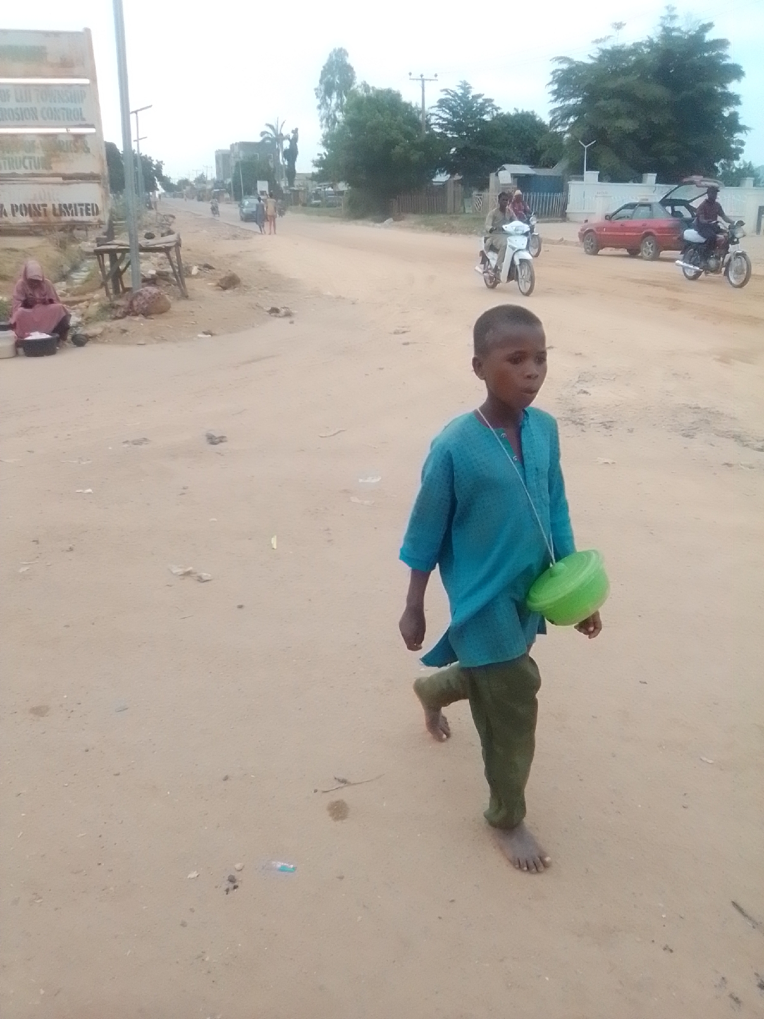 The image depicts a street scene in Gombe, Nigeria. In the foreground, a young boy wearing a teal tunic and green trousers walks barefoot towards the right of the frame. He has a green plastic bowl hanging from a string around his neck. The ground is dusty and unpaved, with scattered debris. In the midground, several motorcycles are visible, some with riders, and a red car with its doors open is parked on the side of the road. Further in the background, buildings and trees line the street. On the left side of the image, a woman in a pink headscarf and garment sits by a roadside stall. A metal signpost with inscriptions is also present on the left. The sky is overcast.
