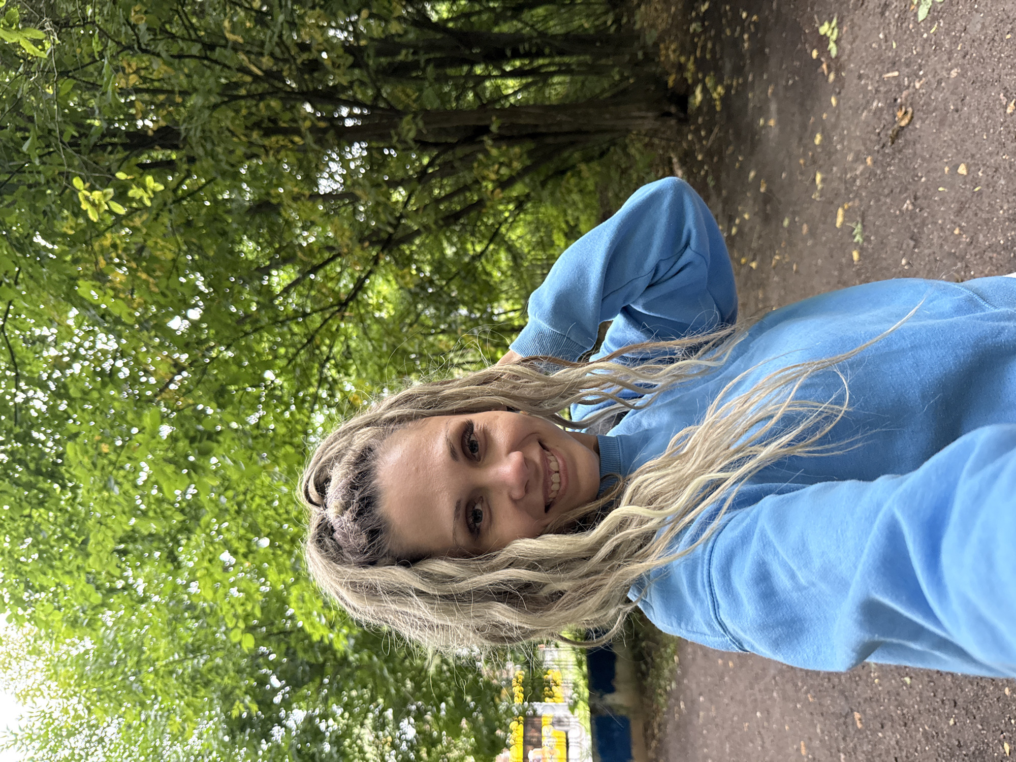 A woman with blonde dreadlocks, styled in a high ponytail with braided sections, smiles at the camera. She is wearing a blue sweatshirt and is outdoors, with trees and foliage in the background. The ground is covered in dirt and fallen leaves. To the left of the woman, there is a structure with colorful painted sections. The overall scene suggests a park or natural setting within the city of Rusanovo, Russia.