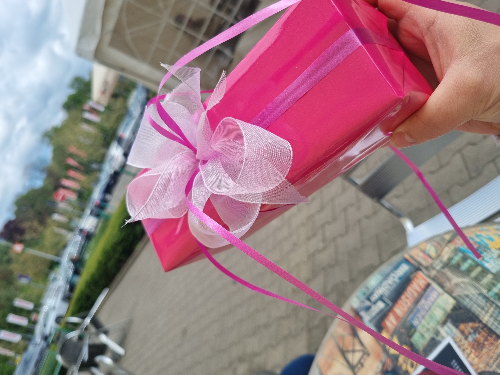 The image presents a tilted, close-up view of a vibrant pink gift box held in a person's right hand. The gift is wrapped in shiny, solid pink paper and adorned with a large, intricate bow made from sheer, light pink organza-like ribbon, complemented by thinner, darker pink satin ribbons that also trail loosely downwards. The hand, with fair skin and short fingernails, grasps the lower right corner of the box.
The background is softly blurred, indicating an outdoor setting during daylight hours, likely under a partially cloudy sky. To the top left, a light-colored building or awning structure is visible. The mid-ground features a line of parked cars, green trees or bushes, and a paved area resembling a street or parking lot. In the bottom right, part of a round table is visible, featuring a distinct patterned surface. This pattern depicts a collage of urban elements, including street signs and cityscapes. Text clearly visible on the table's surface includes "MADISON AV" and "BROADWAY." The ground beneath is paved with light-colored blocks or concrete. No specific activity or emotion is explicitly depicted beyond the act of holding the gift. The scene is set outdoors, possibly at a cafe or public space in Saarbrücken, Germany.