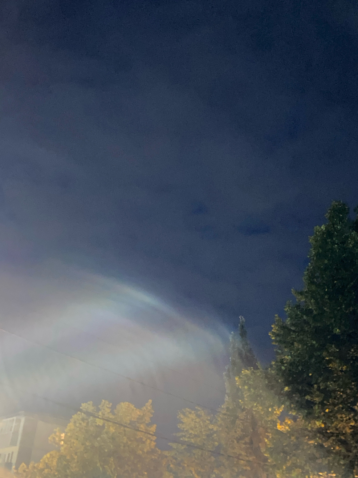 A dark blue sky is visible in the upper portion of the image, with some faint cloud formations. A faint, arc-shaped rainbow is visible in the lower left quadrant, appearing to be refracted light. In the foreground, to the right, is the dark green foliage of a large tree. To the left of this tree, and slightly further back, is a thinner, taller tree with yellow and green leaves. In the far lower left of the image, a portion of a light-colored building is visible. Power lines are visible running horizontally across the lower half of the image. The overall scene suggests an outdoor, nighttime or twilight setting in a city.