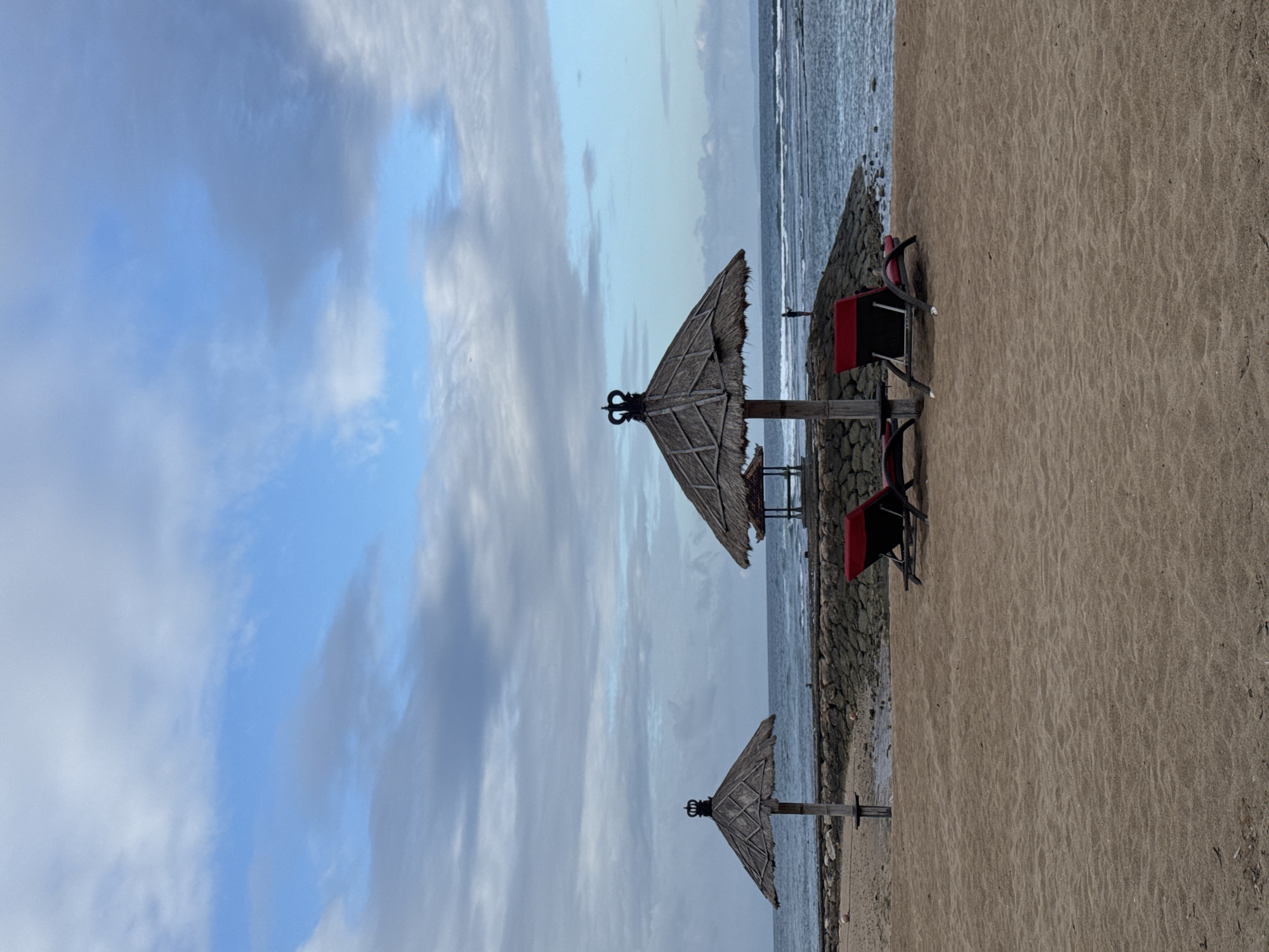 The image depicts a serene beach scene in Kuta Selatan, Indonesia. In the foreground, fine, light brown sand stretches out, marked with gentle ripples. Two red lounge chairs with dark frames are positioned on the sand, facing the ocean. Behind them, a breakwater made of rough, gray stones forms a barrier.
Further back, two thatched-roof umbrellas with decorative crowns on top stand on wooden poles. The umbrella on the left is closer and more prominent, while the one on the right is partially obscured by the breakwater and has a small, open-sided structure behind it. The ocean, a deep blue with white-capped waves, extends to the horizon. The sky is a blend of light blue and muted gray clouds, suggesting either early morning or late afternoon light. In the distance, a faint outline of land can be seen on the horizon. A lone figure, appearing as a small silhouette, stands on the breakwater, looking out at the sea. Two red balls are visible on the sand near the breakwater.