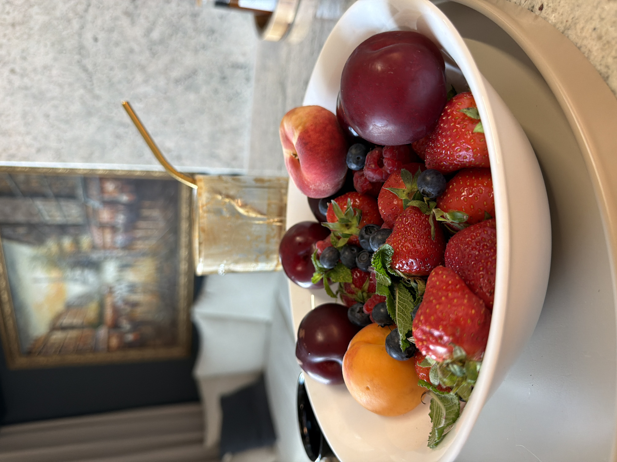 The image shows a white bowl filled with various fruits including plums, strawberries, raspberries, blueberries, a peach, and an apricot. Fresh mint leaves are also visible amongst the fruit. The bowl sits on a tan-colored tray. In the background, out of focus, is a framed painting and a golden straw. The exact location in the city of Plovdiv, Bulgaria cannot be determined from the image. There are no visible people or interactions in the scene.