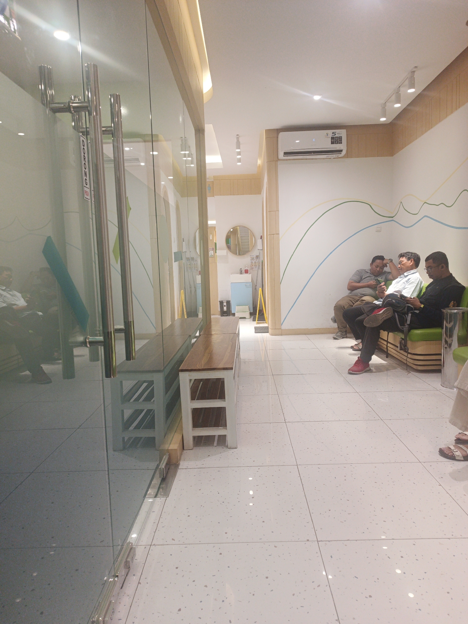 The image depicts a waiting area, likely in a clinic or office building, located in Surabaya, Indonesia. The scene is captured from a slightly elevated angle, looking into a brightly lit interior. In the foreground, a glass door with prominent chrome handles is visible, reflecting the interior of the room. Through the glass, two wooden benches with white bases are positioned, one longer than the other, with a shelf underneath the longer bench.
The main focus of the scene is a group of four men seated on a bright green sofa against a white wall adorned with wavy, multi-colored lines in green, yellow, and blue. The men appear to be waiting. The man on the far left is wearing a grey shirt and dark pants, looking at his phone. Next to him, a man in a light blue collared shirt and dark pants is also engrossed in his phone. The third man from the left, in a grey polo shirt and brown pants, rests his head on his hand and looks down. The man on the far right is wearing a black shirt and dark pants, also looking at his phone. All men are wearing shoes, with one man on the left wearing red sneakers.
In the background, an air conditioning unit is mounted high on the wall. Further down the waiting area, a circular mirror hangs on the wall, and to its left, a small blue cabinet with a white sink is visible. A yellow stand with a sign is positioned near the cabinet. The floor is tiled with large, light-colored square tiles with a speckled pattern. The overall ambiance suggests a clean, modern, and well-maintained waiting space.