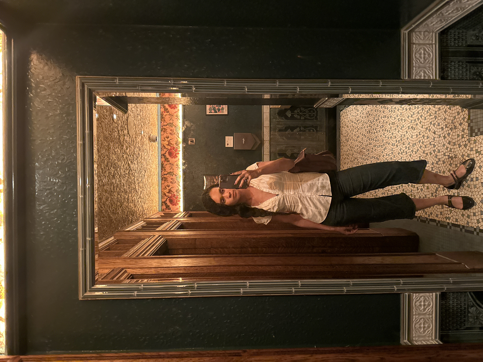 A woman is seen reflected in a mirror within an enclosed space. The woman is taking a selfie with her phone, holding it in her right hand. She is wearing a white, button-down shirt and dark pants, with dark shoes. A brown bag rests near her. The reflection reveals the interior of what appears to be a room with distinct decor, including textured wallpaper, mosaic flooring, and patterned wall panels. Details visible in the reflection, such as the patterned wallpaper, the mosaic floor, and specific wall panels, strongly suggest the location is the restroom at the Brasserie Zedel in London, United Kingdom. The image also includes the surrounding ornate framing of the mirror and textured dark green walls outside the mirror's reflection.