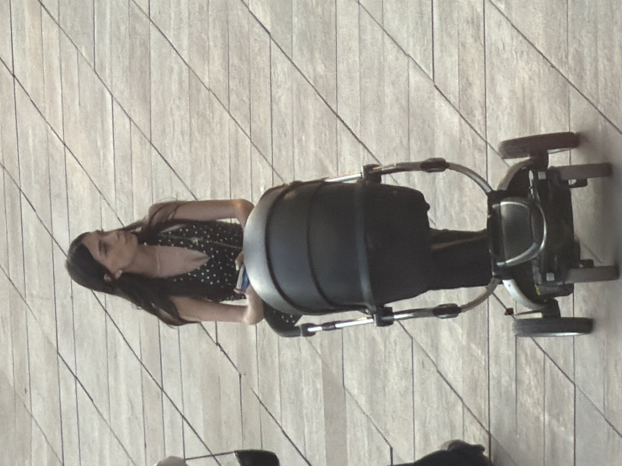 In a high-angle shot, a young woman with long, dark hair is captured from the side, pushing a black stroller across a light-colored wooden floor. She is wearing a black dress with white polka dots. Her gaze is directed downwards, her expression unreadable. The stroller, a prominent object in the scene, features a large, rounded, black main body and a metal frame with wheels. The floor, made of thin wooden planks laid diagonally, creates a textured backdrop. The lighting suggests it is daytime, possibly indoors, with the light source appearing to come from above and to the left. No text is visible in the image. The setting appears to be a public indoor space, perhaps a shopping mall or an exhibition hall, given the polished wooden floor and the presence of the stroller. The scene is static, focusing on the woman and the stroller, with no other individuals or distinct environmental features visible. The overall mood is neutral, with the woman's downward gaze offering a slight hint of introspection or focus on her task.
