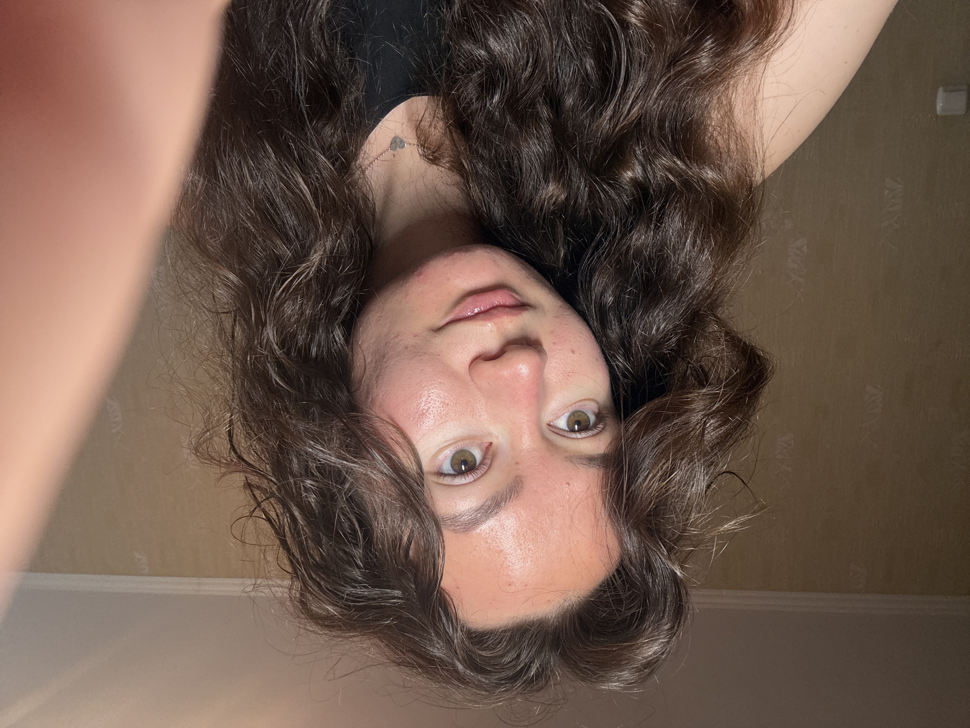 A selfie of a young woman with wavy brown hair and fair skin. She is looking directly at the camera with a slight upward gaze. Her eyes are hazel, and her lips are glossy. She is wearing a black top and a delicate necklace. The background is a beige wall with a faint pattern. The location, Chaikovsky, Russia, cannot be verified from the image.