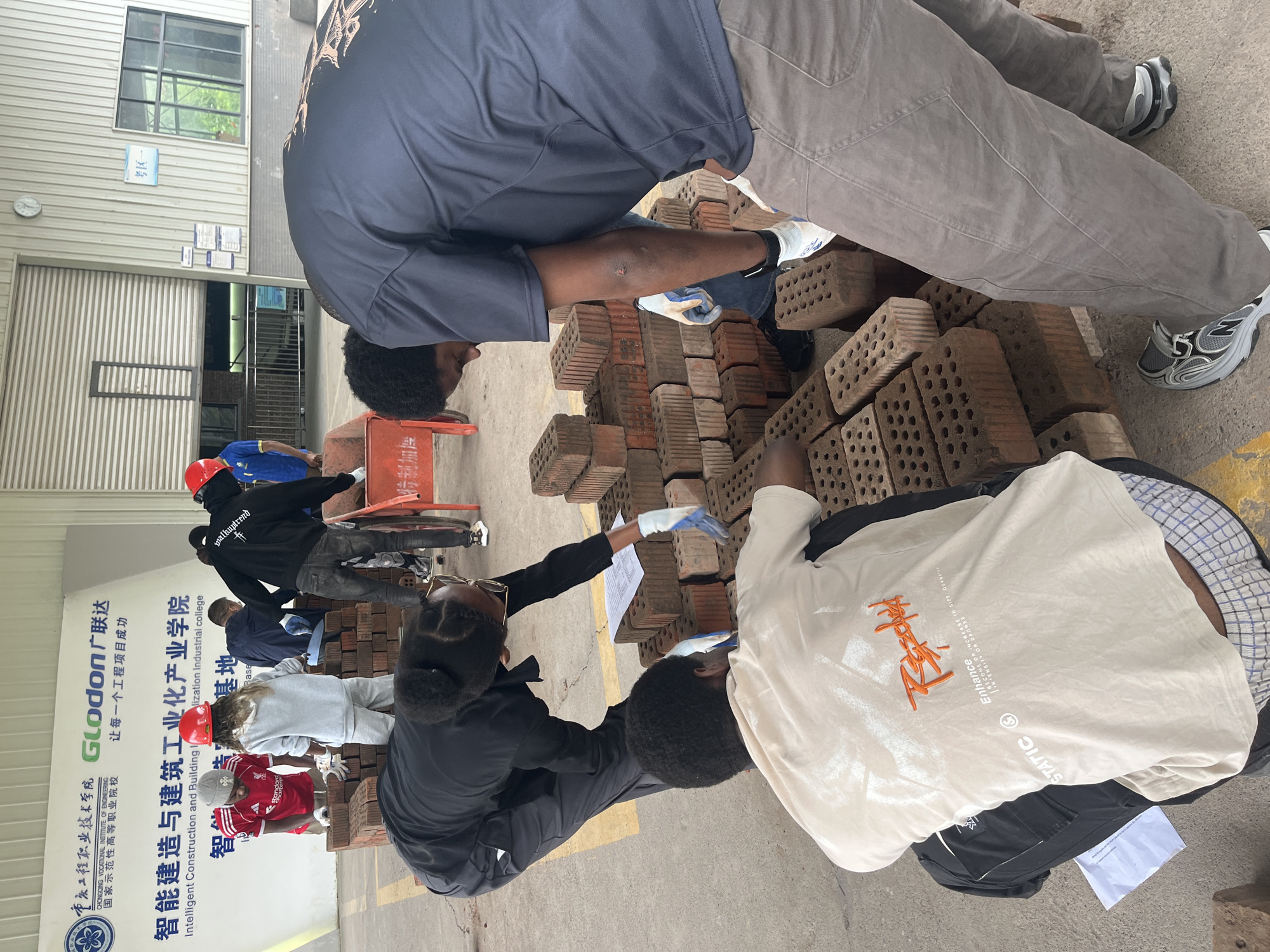 The image depicts an outdoor vocational training environment, likely at the Intelligent Construction and Building Industrialization College in Jiangjin, China. The foreground shows several individuals engaged in bricklaying activities.
In the center, a person wearing a black hoodie and dark pants, with a red hard hat, is actively working with bricks. To their left, another individual, dressed in a grey hoodie and lighter pants, also wearing a red hard hat, is interacting with the brick pile. A third person, in a black long-sleeved shirt and dark pants, is leaning over a stack of bricks, possibly inspecting them or preparing to lay them. They are wearing white gloves. Another individual, wearing a light-colored t-shirt with graphic text and checkered shorts, is seated amongst the bricks, looking down at the ground. Their t-shirt has the words "Enhance" and "STATIC" printed on it.
In the background, a large banner is visible with Chinese and English text, indicating the institution's name and educational focus. The environment appears to be an open-air workshop or training ground, with concrete flooring and industrial building structures visible. Stacks of bricks are prominently featured throughout the scene. A red wheelbarrow is also present, further suggesting construction or training activities. The lighting suggests it is daytime.