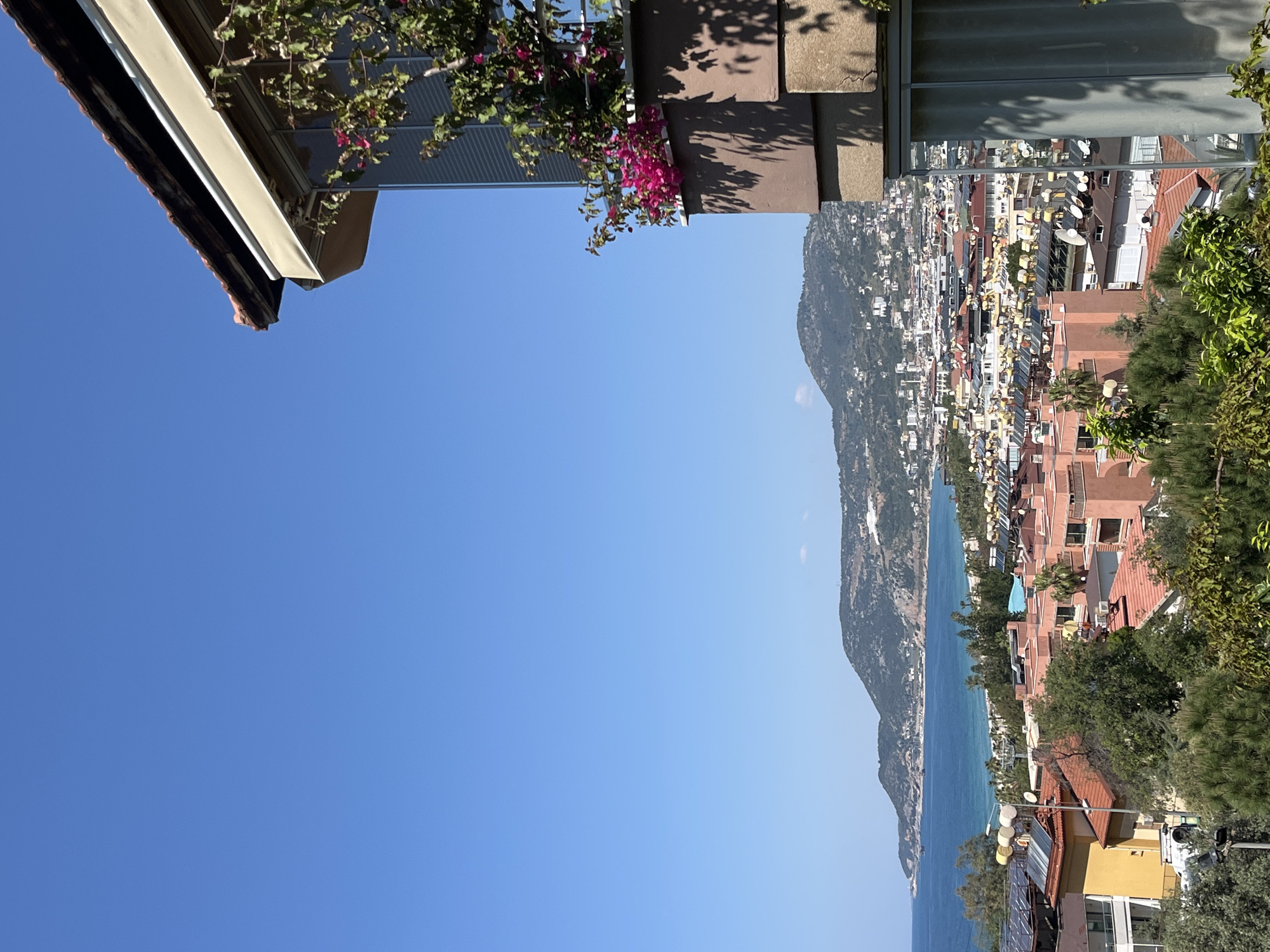 The image captures a view from an elevated position overlooking Alanya, Turkey. The foreground features architectural elements, including a building roof and a balcony adorned with foliage and pink flowers. In the midground, the urban landscape of Alanya stretches along the coastline, comprised of closely packed buildings of varying heights, predominantly with red-tiled roofs and light-colored facades. A mountain range forms a backdrop, creating a distinct separation between the city and the sky. The Mediterranean Sea is visible as a turquoise-blue strip along the edge of the city. The clear, bright blue sky dominates the upper portion of the image.