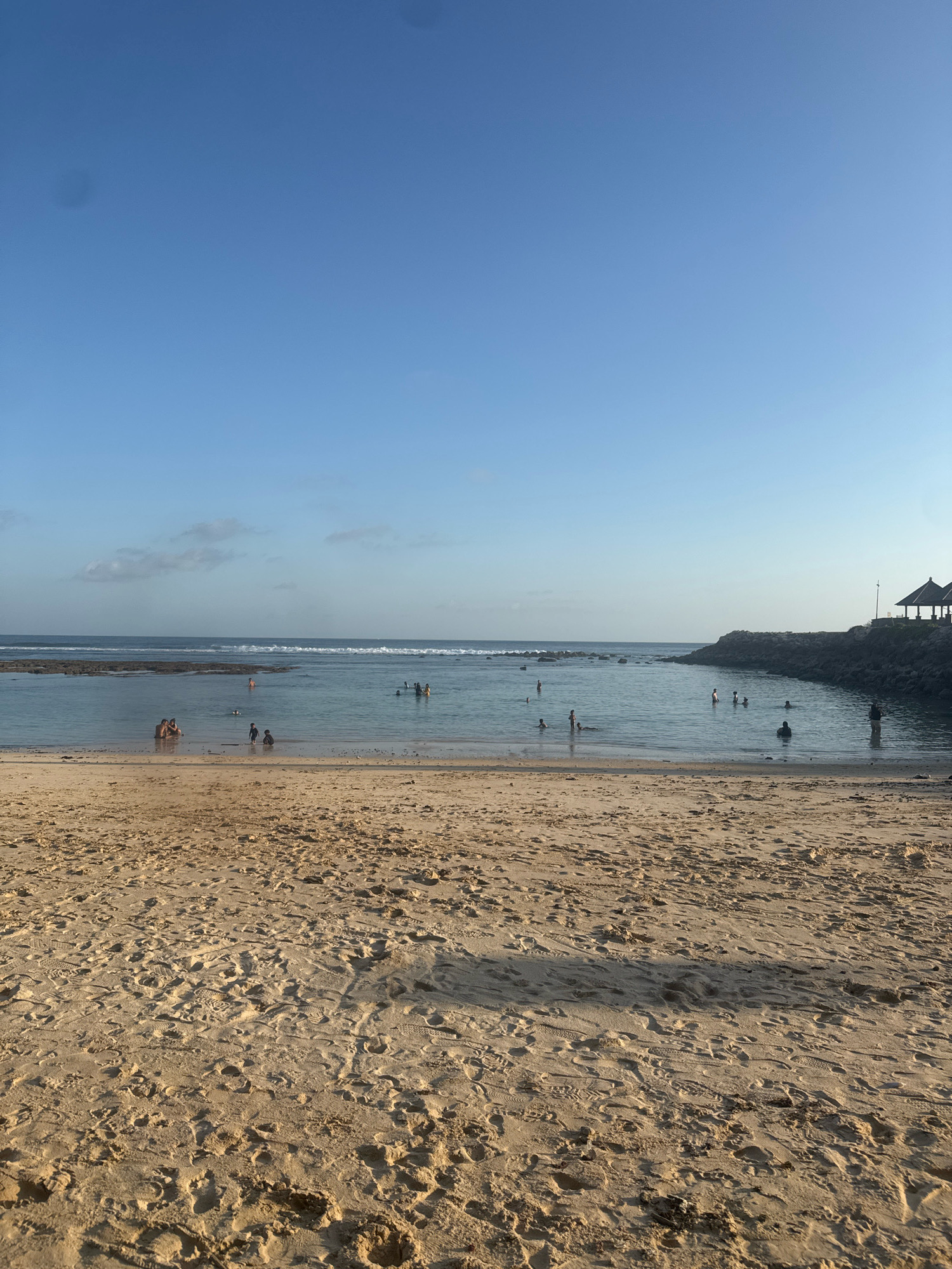 A serene beach scene unfolds under a clear blue sky in Kuta Selatan, Indonesia. The foreground is dominated by a stretch of sun-kissed sand, marked with numerous footprints and the gentle indentations of rippled sand. Beyond the sand, the tranquil ocean stretches out, its surface calm with gentle waves approaching the shore.
Scattered across the shallow water are various individuals engaged in recreational activities. Some are wading, while others are swimming. A small group of people are gathered near the shore, seemingly interacting. In the distance, a rocky breakwater extends into the sea, creating a sheltered cove. Atop this breakwater, a traditional Balinese-style gazebo with a thatched roof offers a shaded respite, overlooking the ocean. The overall impression is one of a peaceful and inviting coastal environment, characteristic of Kuta Selatan.