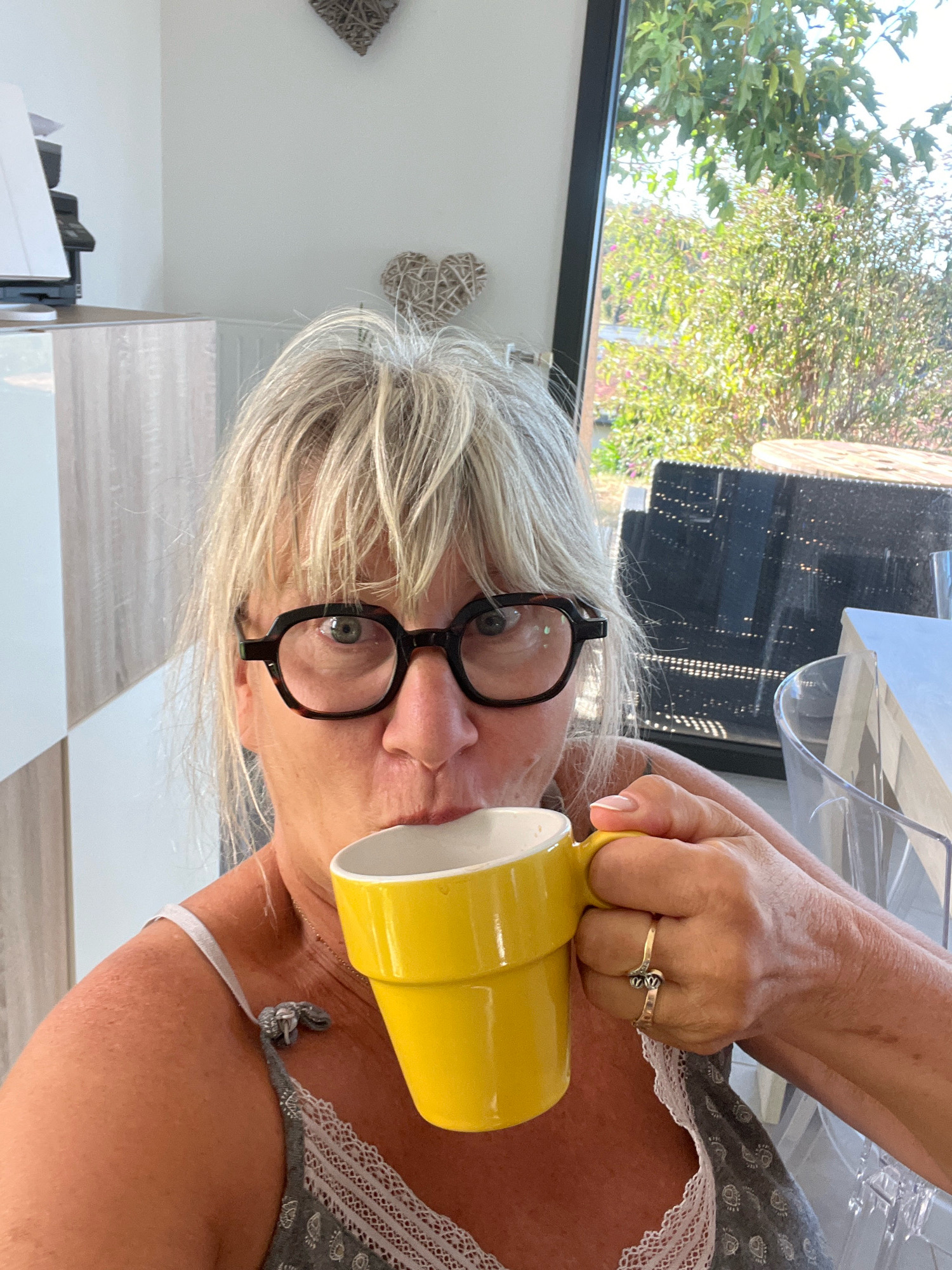 A woman with blonde hair and glasses is drinking from a yellow mug. She is wearing a gray and white top. The scene appears to be indoors, possibly in a kitchen, with a window in the background showing greenery and trees. There is a desk with a printer to the left of the image. The location is identified as Chamboeuf, France.