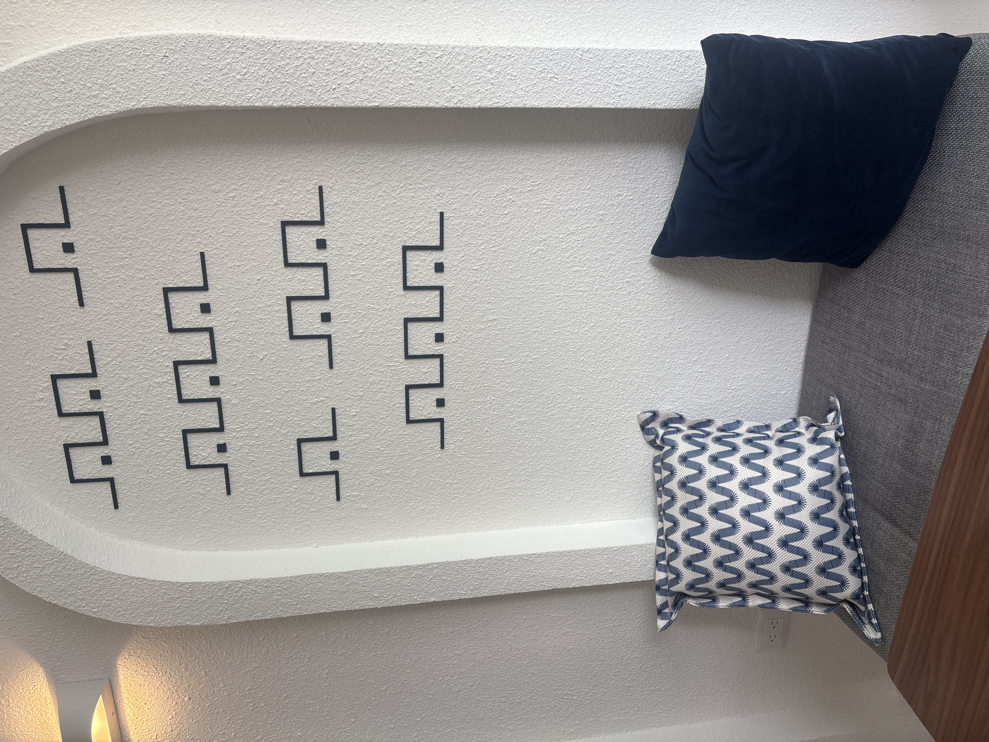 The image shows a white, textured wall in an interior space. The wall features a decorative arched recess containing a series of stylized, geometric black line drawings. There are two throw pillows placed against the wall: a dark blue velvet pillow and a smaller, blue and white patterned pillow. An electrical outlet is visible near the smaller pillow. A portion of a gray upholstered piece of furniture with a wood accent panel is seen to the right. This image was most likely taken in a private home. Location of the city of Puebla, Mexico, is unconfirmed.